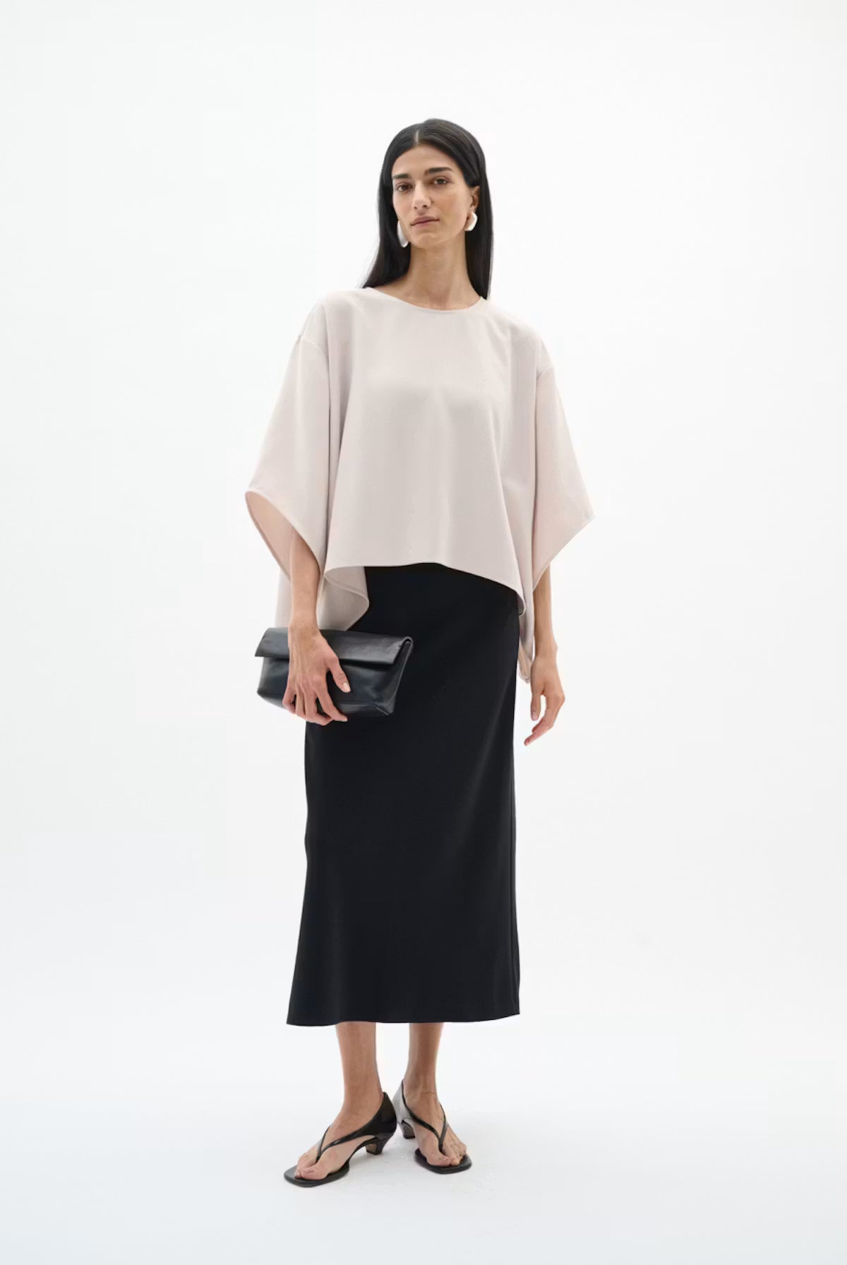 A woman modeling a light blush pink draped short-sleeve top and a black midi skirt, holding a black clutch, against a plain white studio background.