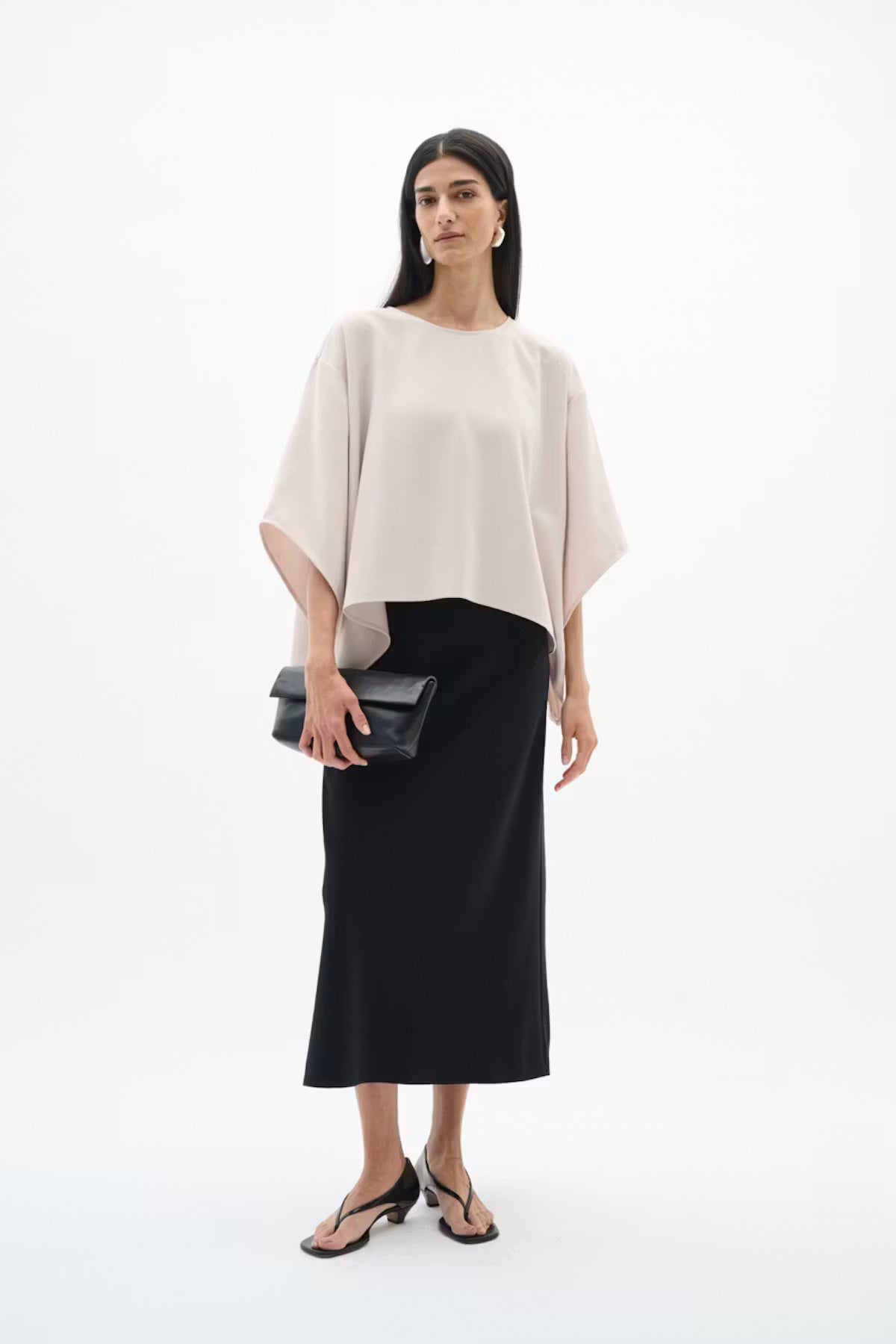 A woman modeling a light blush pink draped short-sleeve top and a black midi skirt, holding a black clutch, against a plain white studio background.