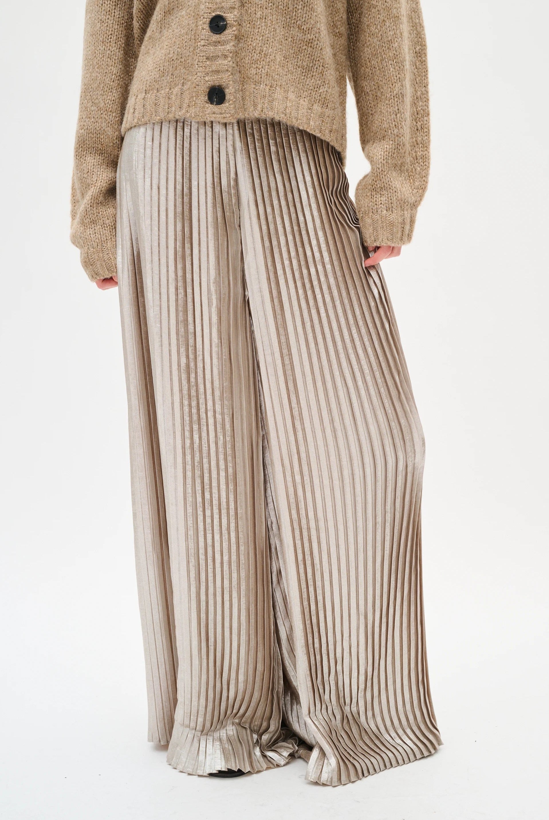 Person wearing a beige cardigan and silver pleated pants on a white background