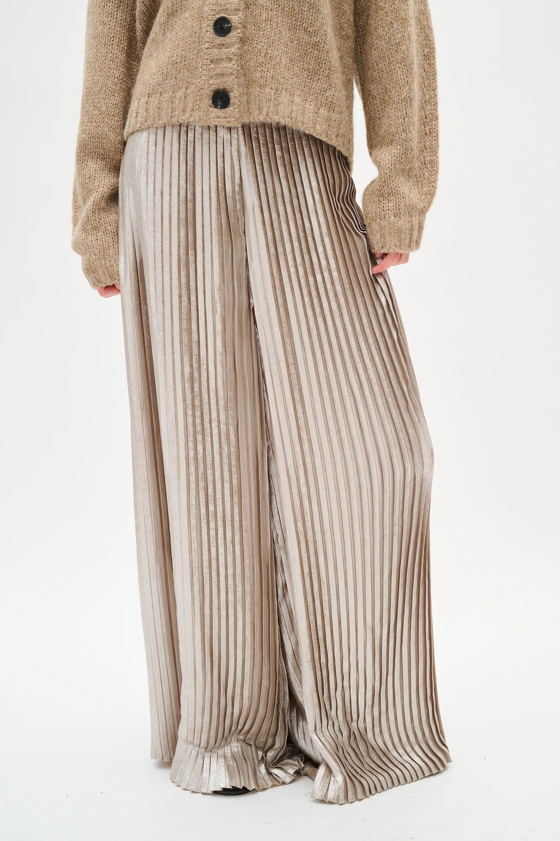 Person wearing a beige cardigan and silver pleated pants on a white background
