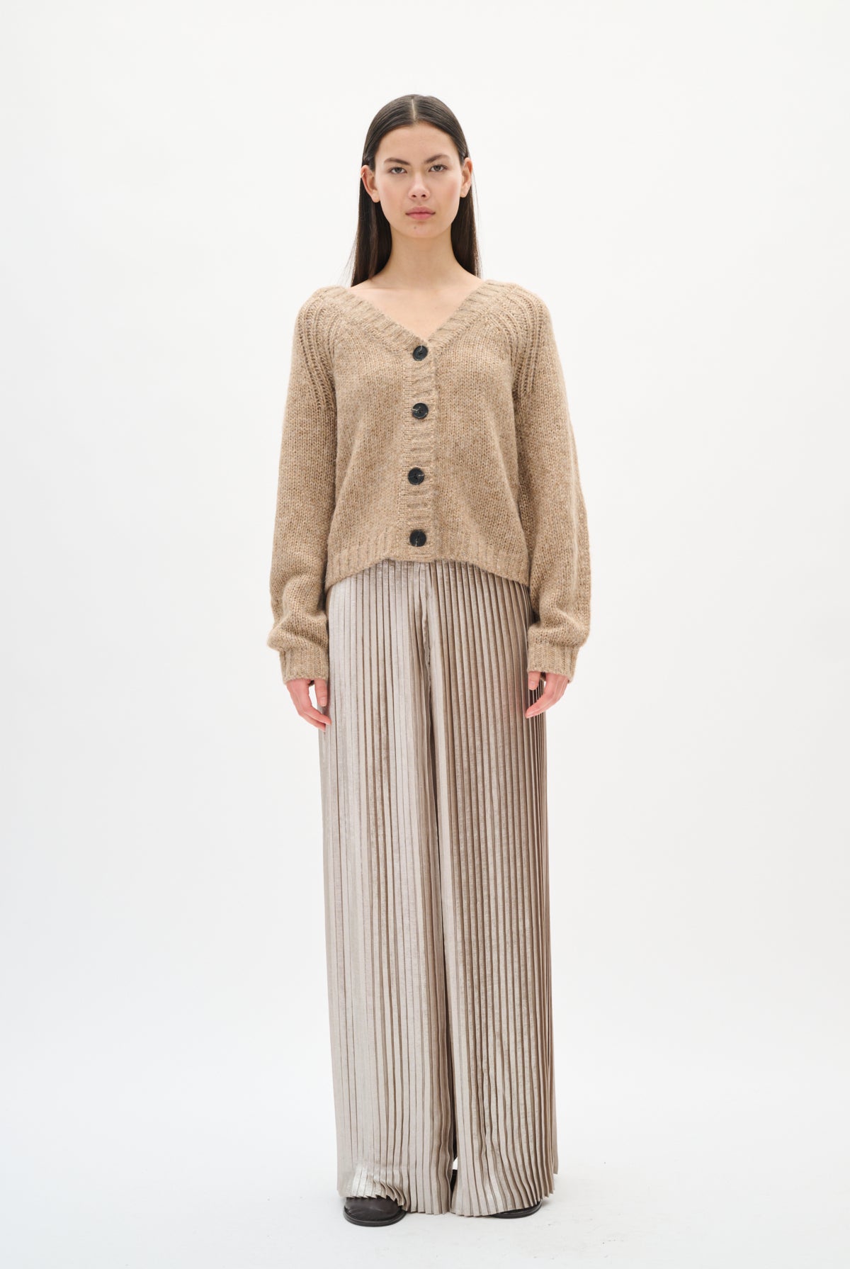 Person wearing a beige cardigan and silver pleated pants on a white background