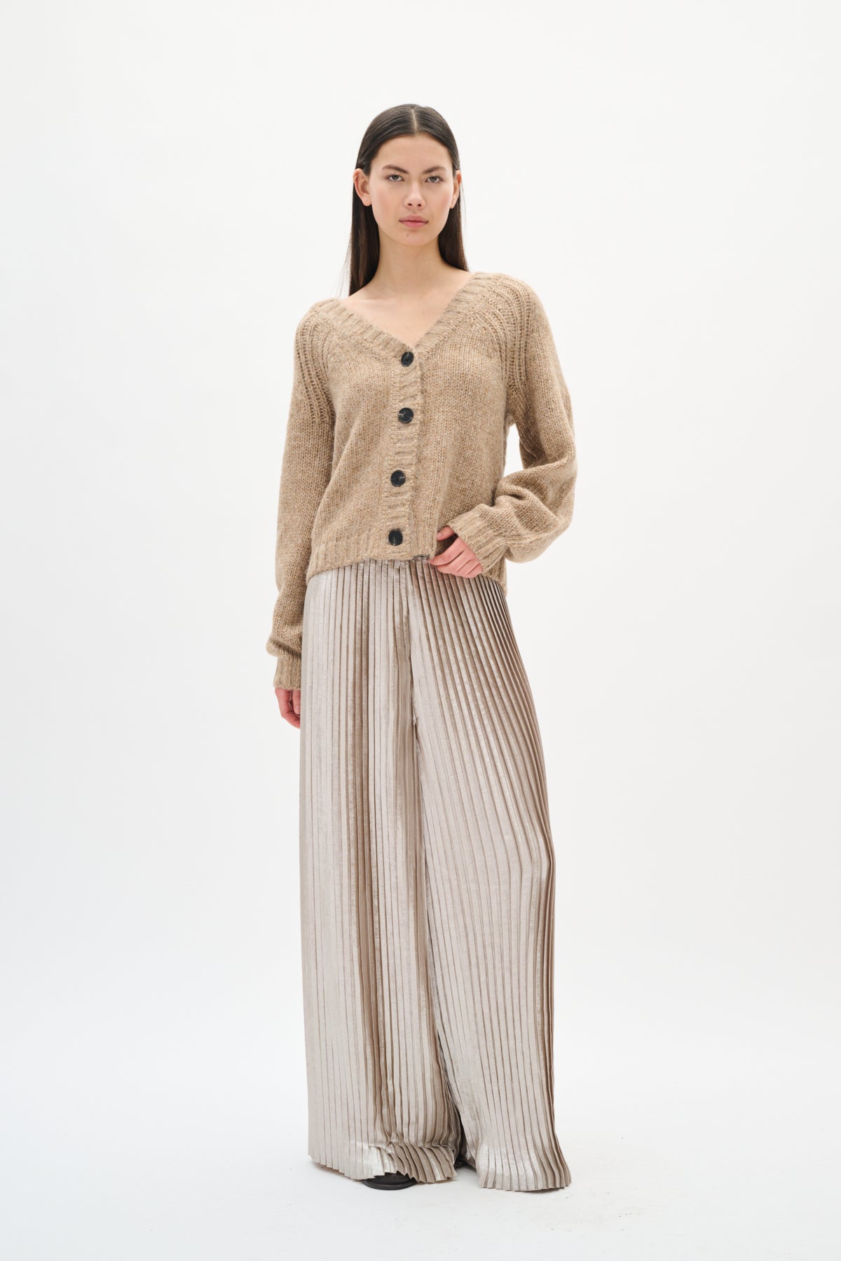 Person wearing a beige cardigan and silver pleated pants on a white background