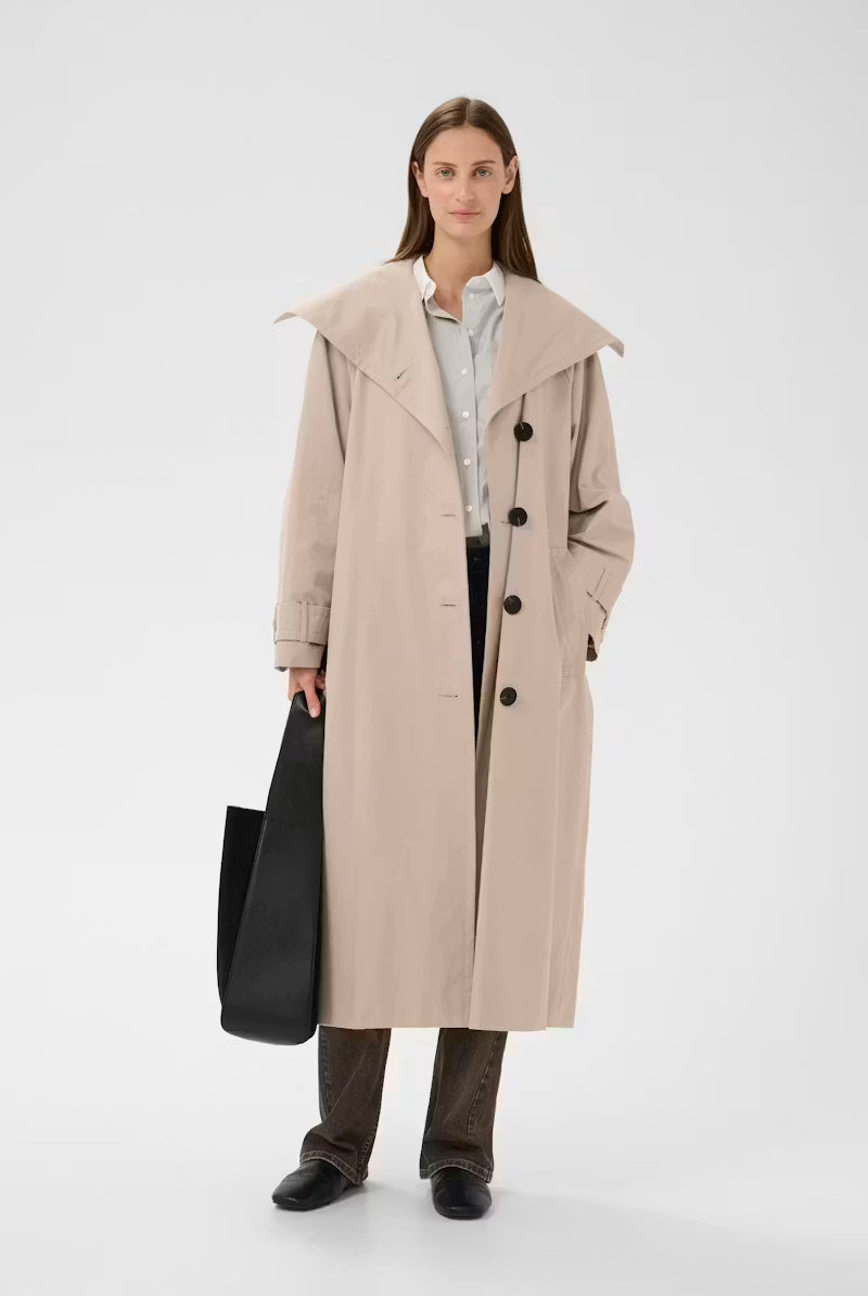 A woman stands against a light gray studio background wearing a long beige trench coat over a white shirt and dark pants, while holding a large black tote bag.
