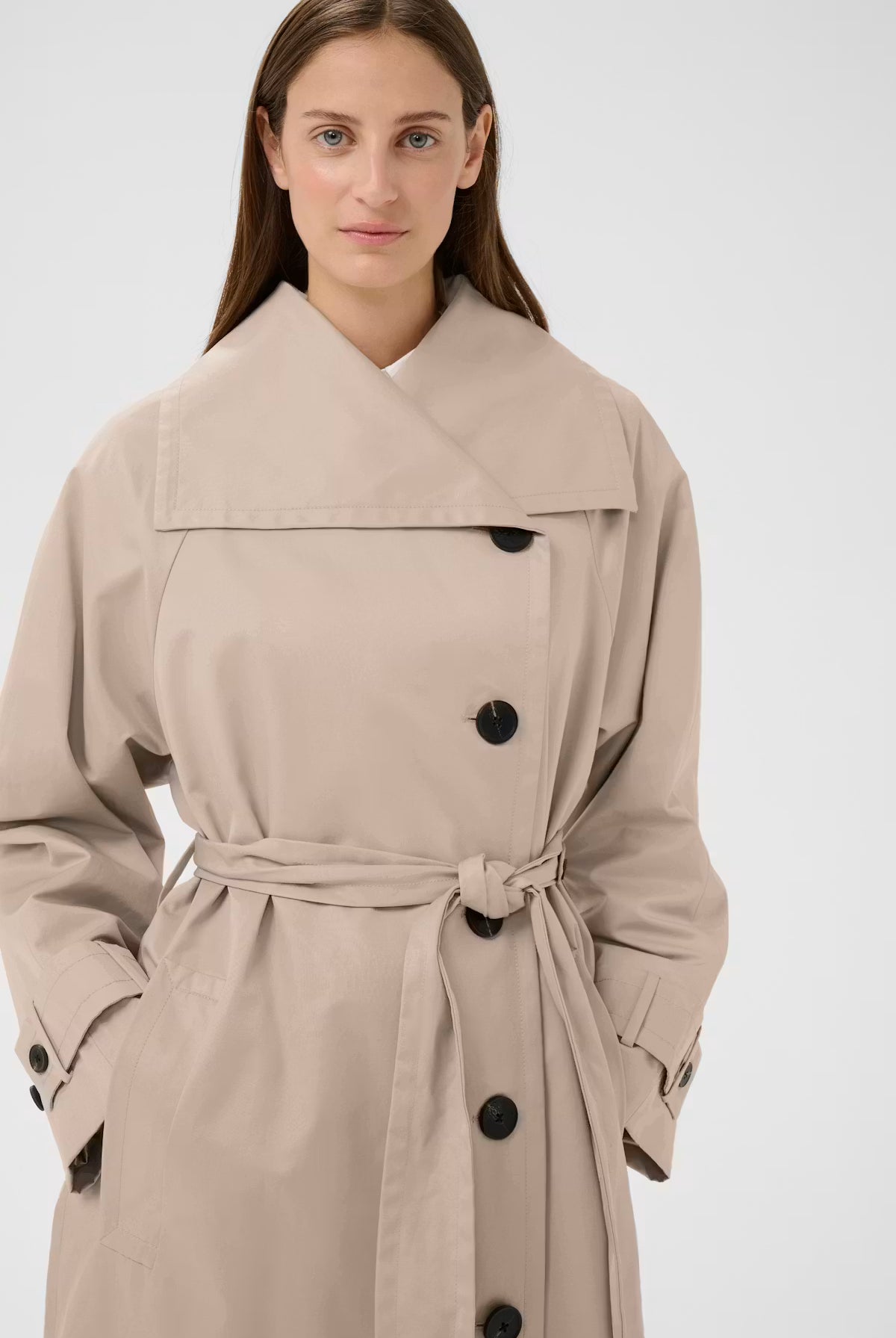 A woman wearing a beige trench coat with a large collar, black buttons, and a matching waist tie, photographed against a plain light gray studio background.