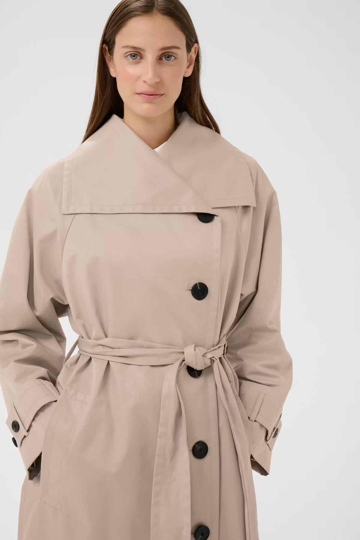 A woman wearing a beige trench coat with a large collar, black buttons, and a matching waist tie, photographed against a plain light gray studio background.