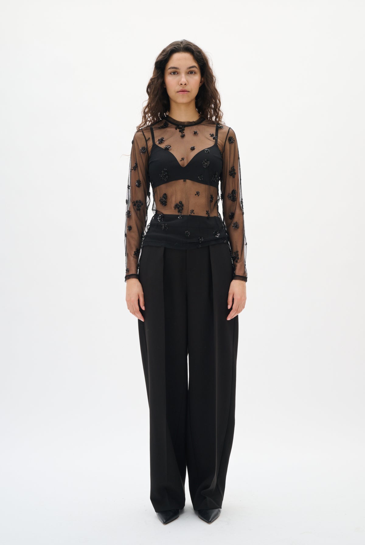 Model wearing a sheer black top with floral patterns and high-waisted black pants on a white background