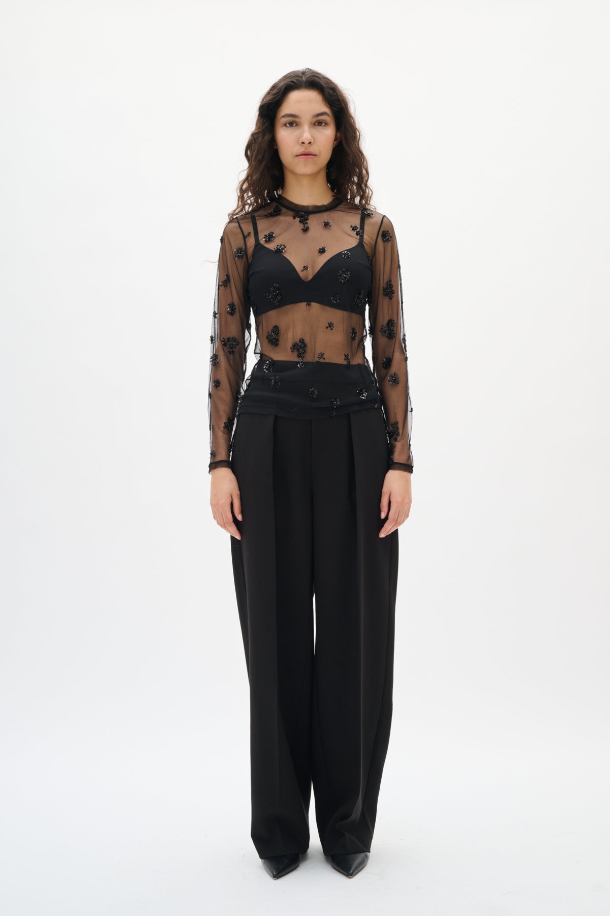 Model wearing a sheer black top with floral patterns and high-waisted black pants on a white background