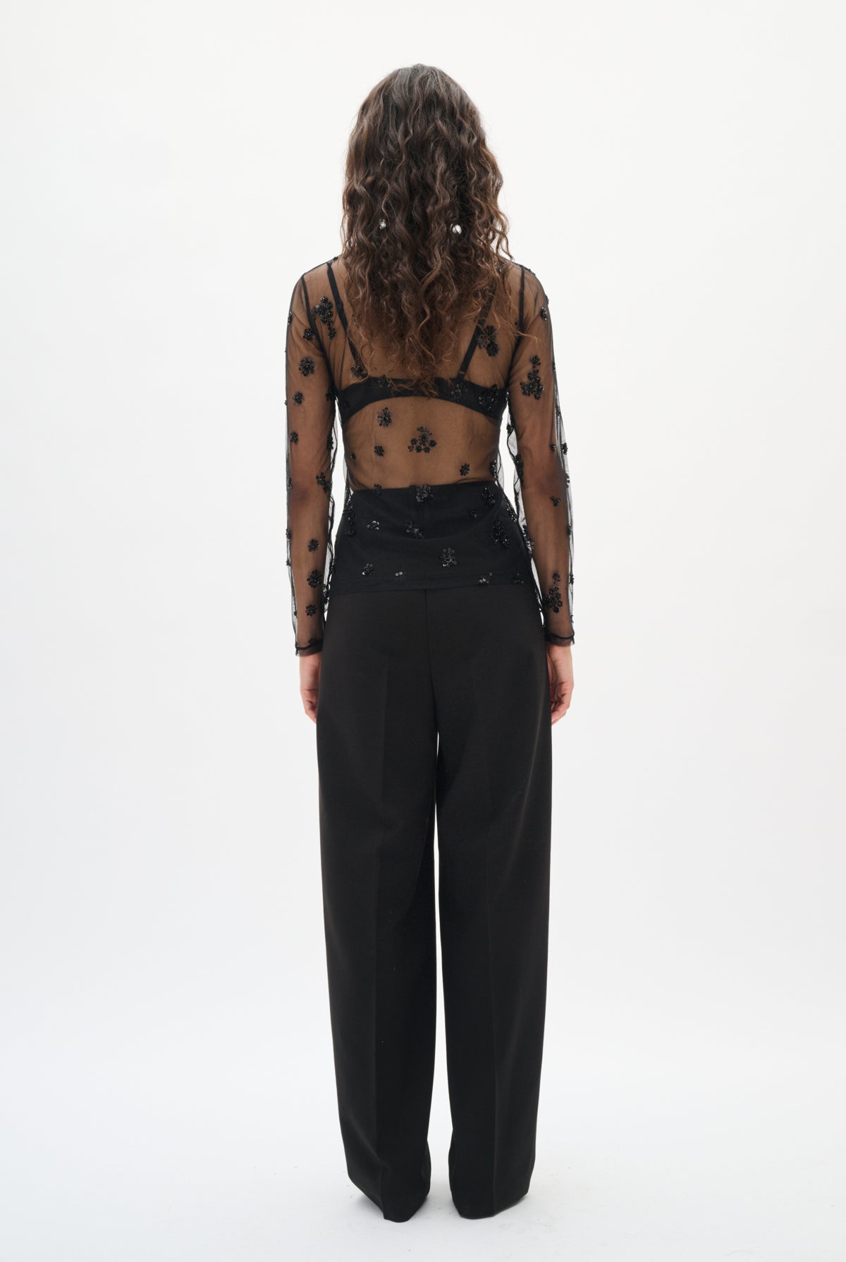 Person wearing a sheer top with black polka dots and high-waisted black pants on a white background back angle