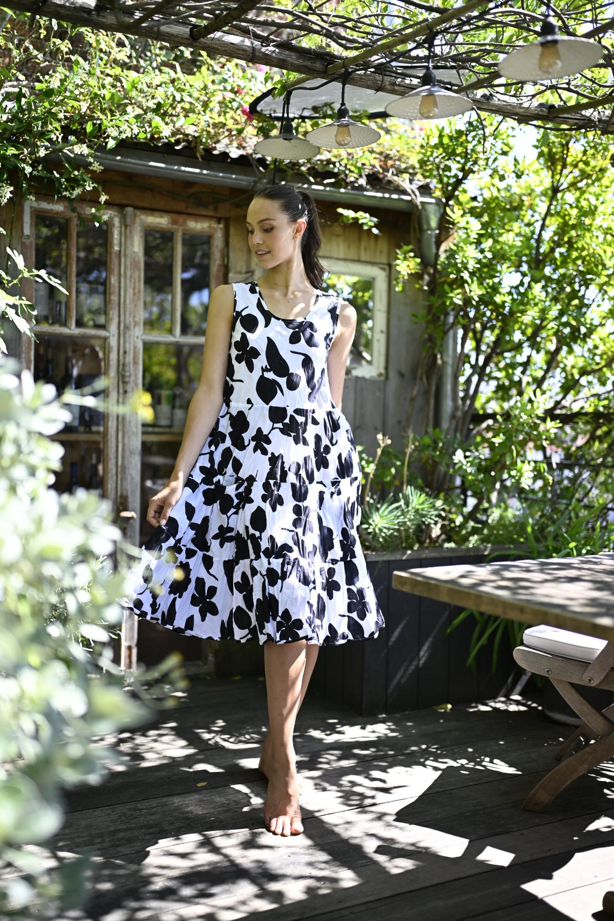 A woman wearing a sleeveless black and white floral print dress stands barefoot on a wooden deck in a garden setting with a rustic cabin and pergola in the background.