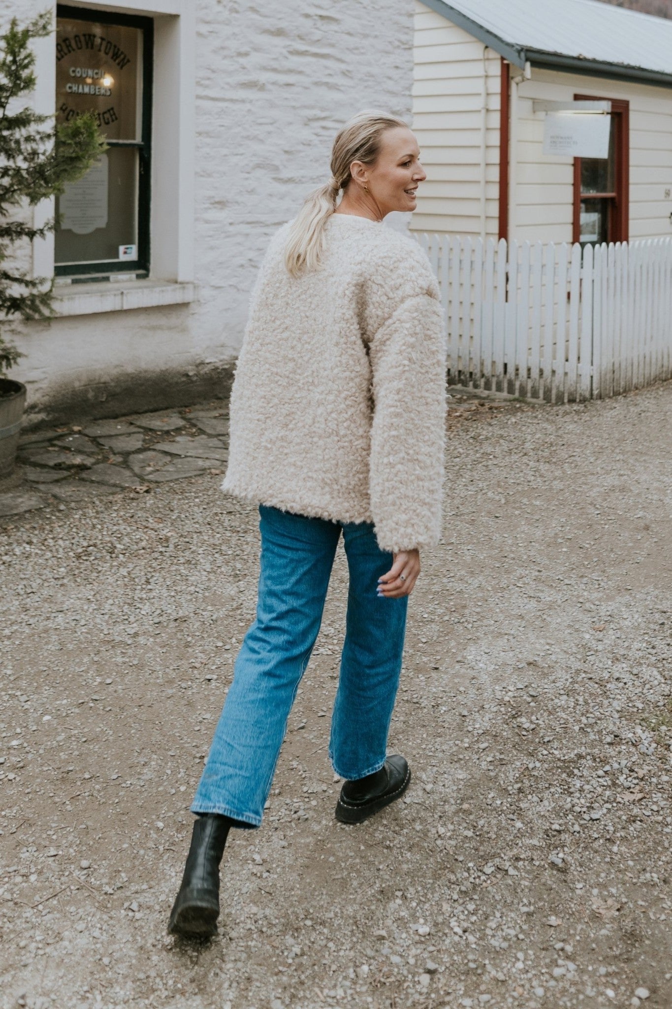 Woman wearing a cream shearling jacket and blue jeans standing on a gravel driveway.