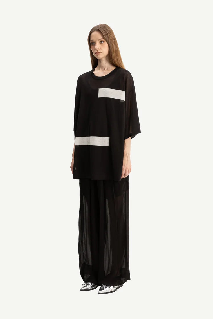 A model wearing a black wide-sleeve top with two horizontal white stripes and matching black wide-leg sheer pants, standing against a solid off-white background.