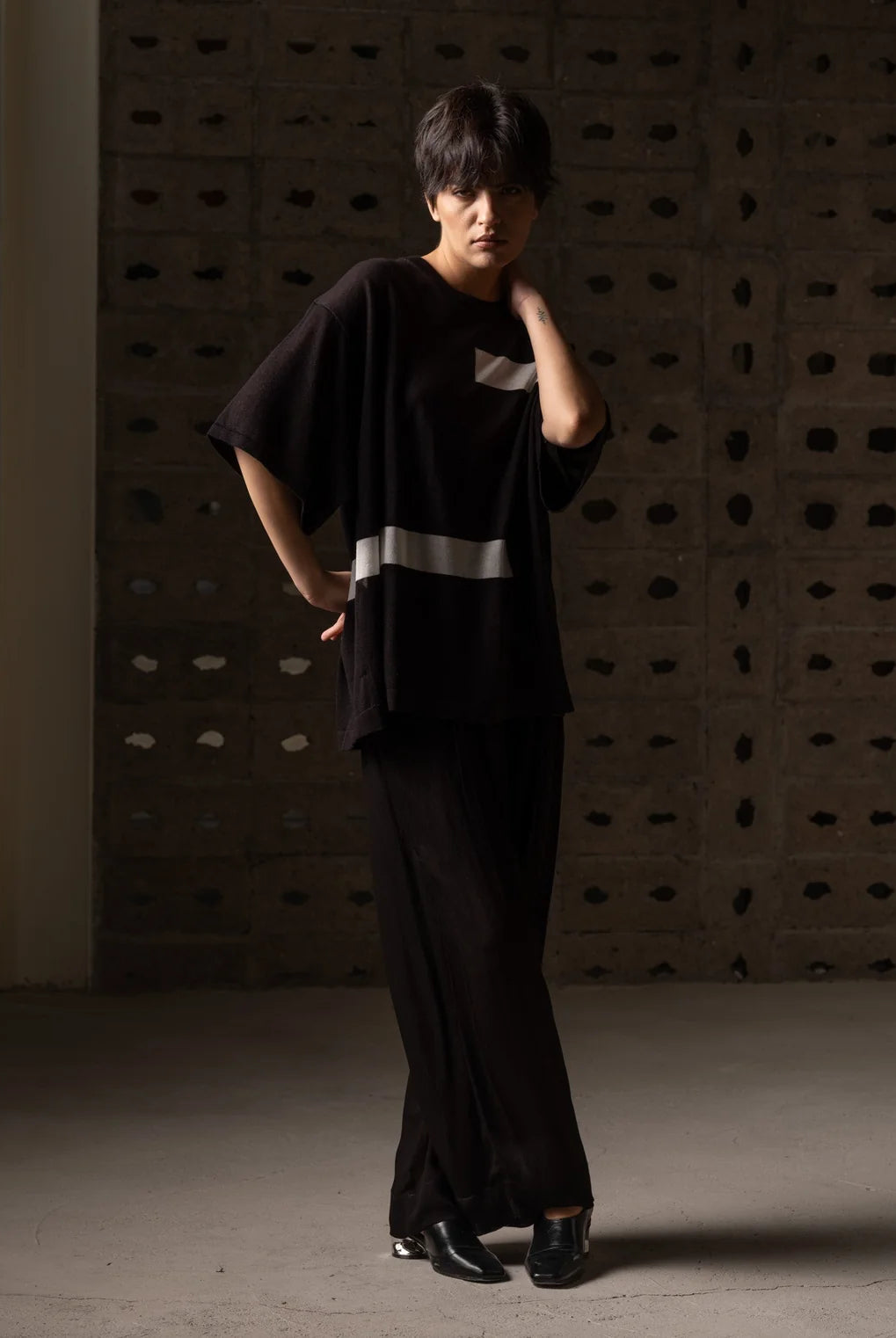 A model wearing a black oversized short-sleeve top with a silver reflective stripe and matching black wide-leg trousers, standing in front of a textured concrete wall.