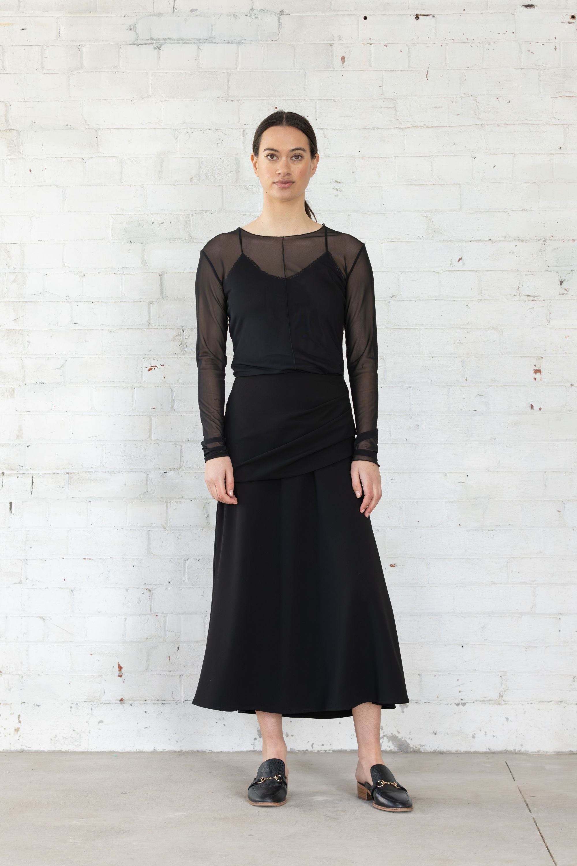 Woman wearing a black dress with sheer sleeves against a white brick wall.