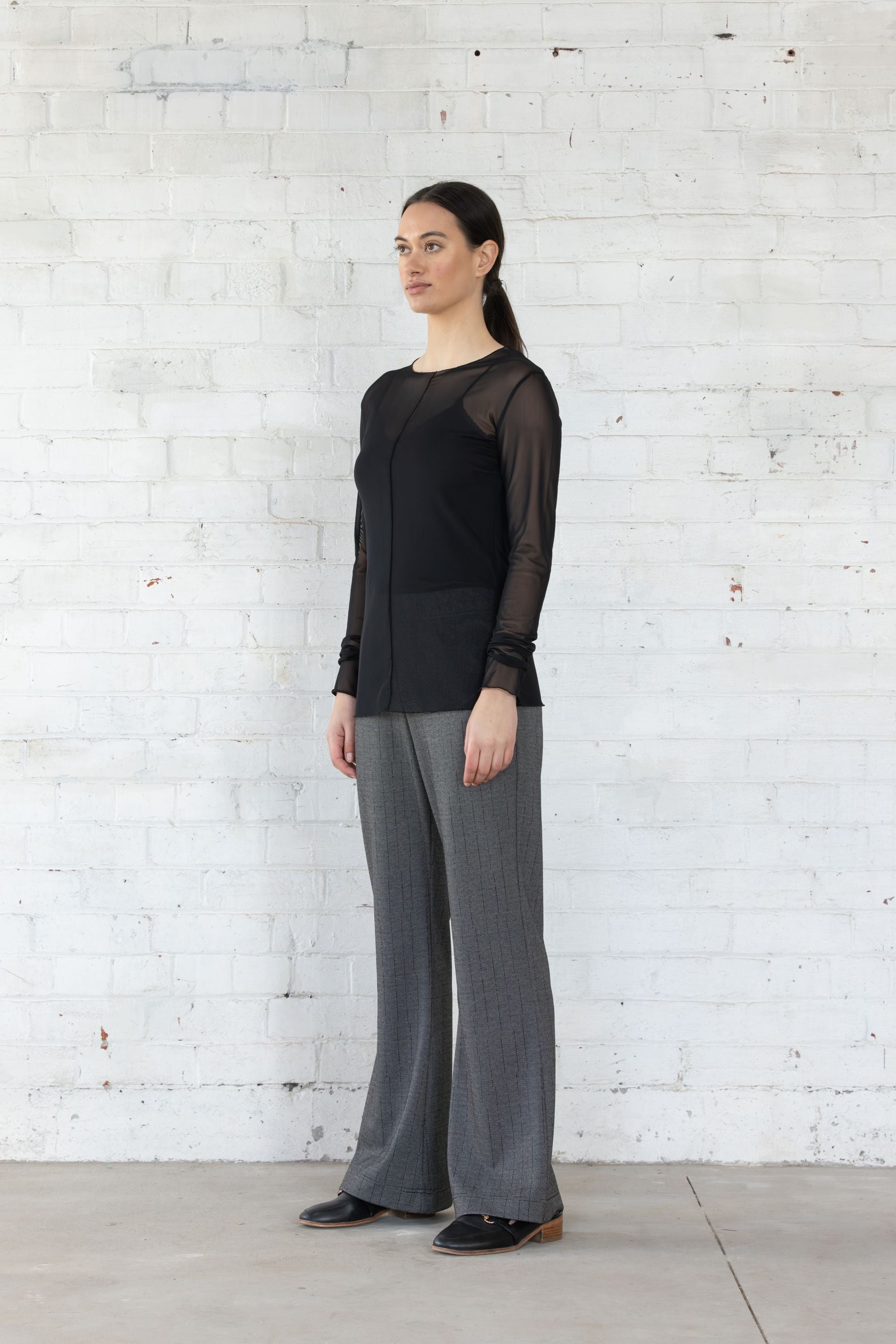 Woman wearing a black top and gray pants standing against a white brick wall.