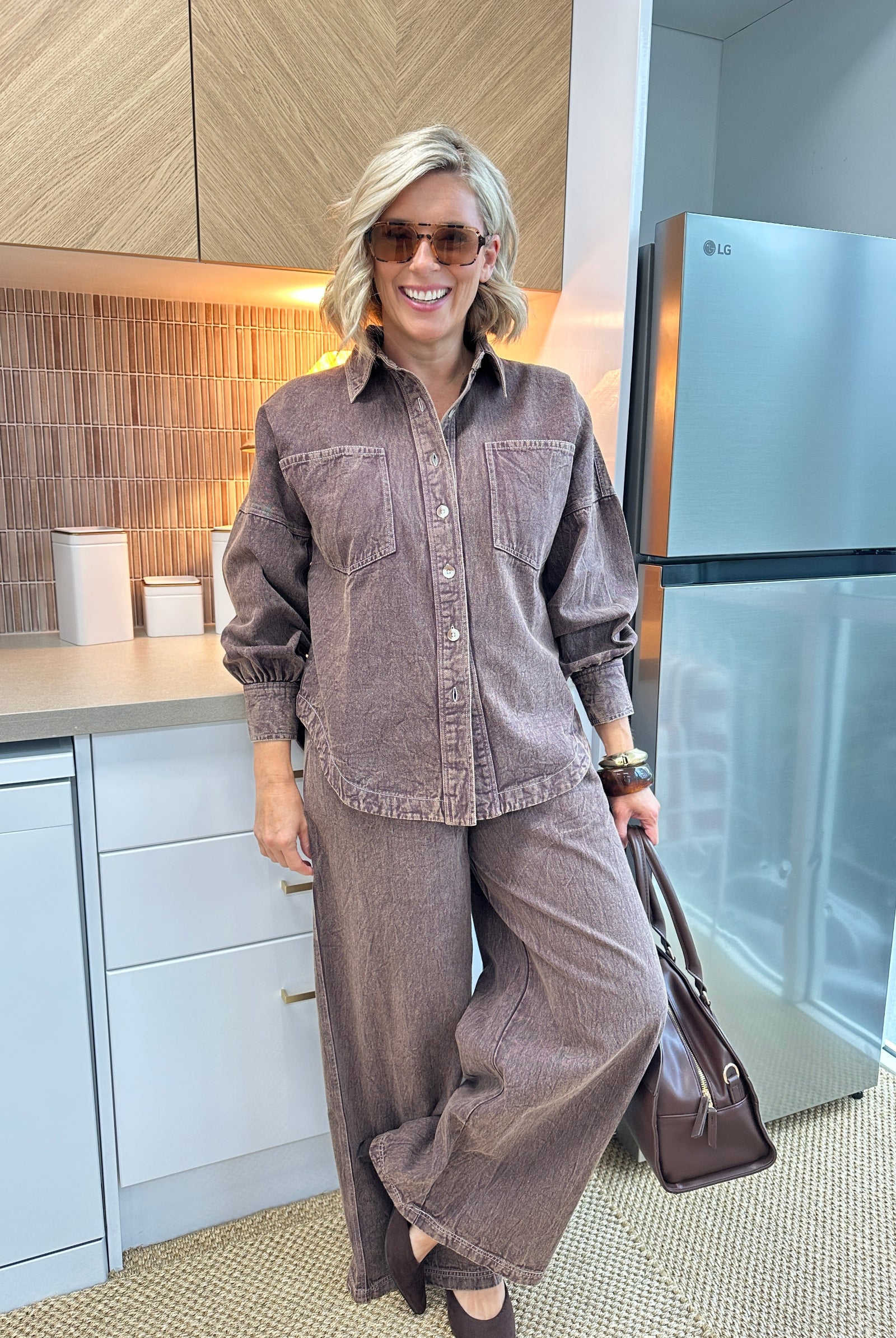 A woman wearing a matching brown corduroy button-up shirt and wide-leg pants set, holding a brown leather handbag in a modern kitchen setting.