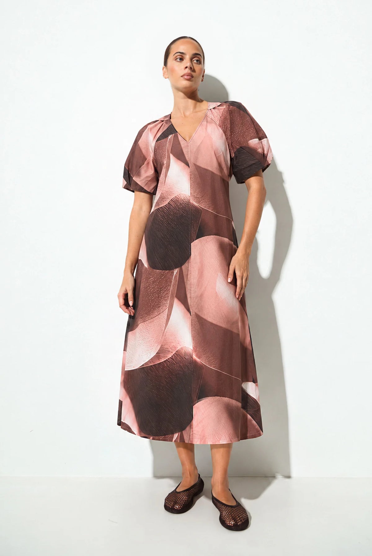 A woman modeling a midi-length dress with a pink, brown, and beige abstract floral pattern, featuring short puff sleeves and a V-neckline, set against a plain white studio background.