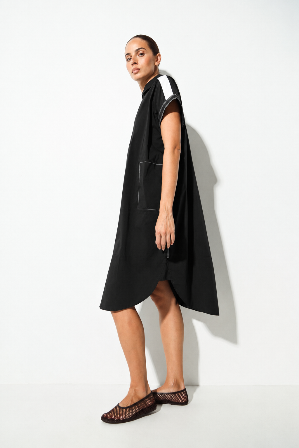 A woman modeling a black sleeveless midi dress with a high collar, a large front patch pocket, and contrasting white stripe details on the shoulders, set against a plain white studio background.
