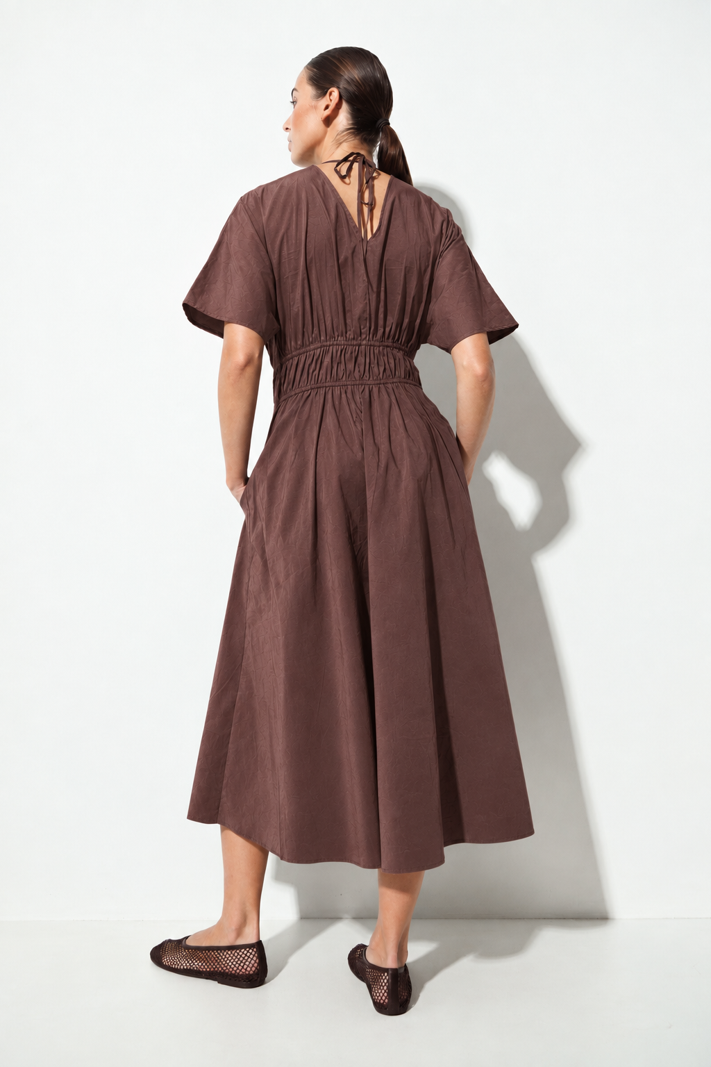 A woman wearing a brown short-sleeve midi dress with a smocked waist and tie-back neckline, shown from the back against a white studio background.