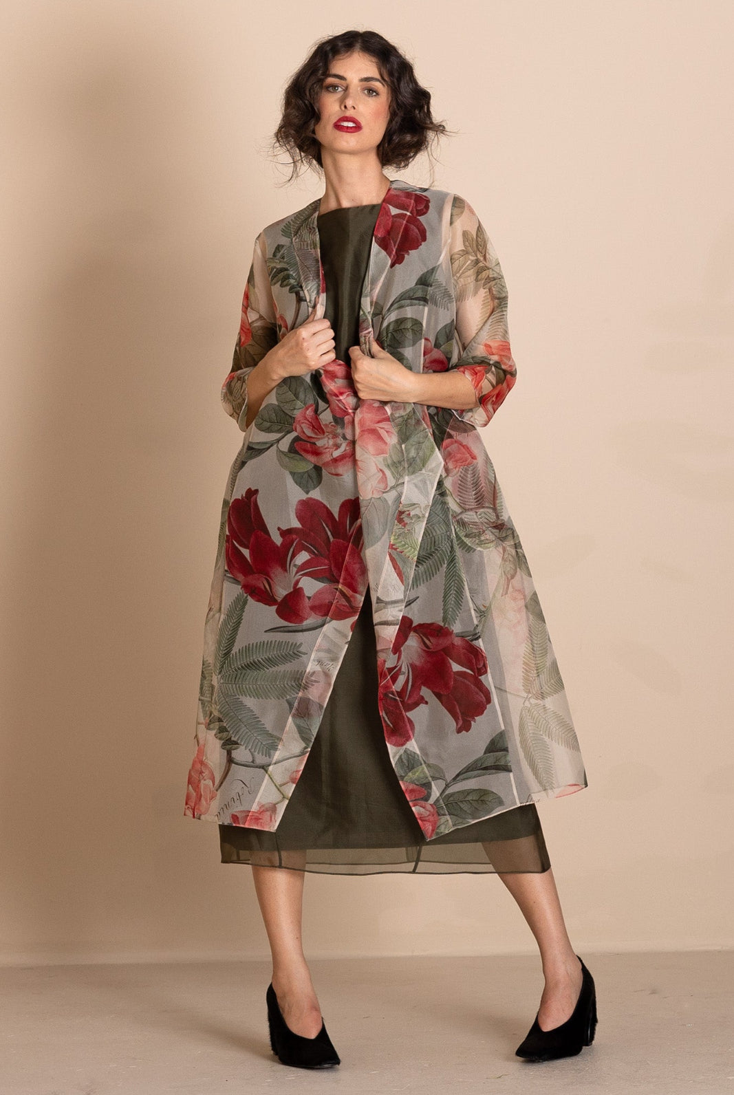 A woman models a sheer, lightweight open-front duster jacket featuring a large red and green floral print over a dark olive green dress, set against a solid beige studio background.