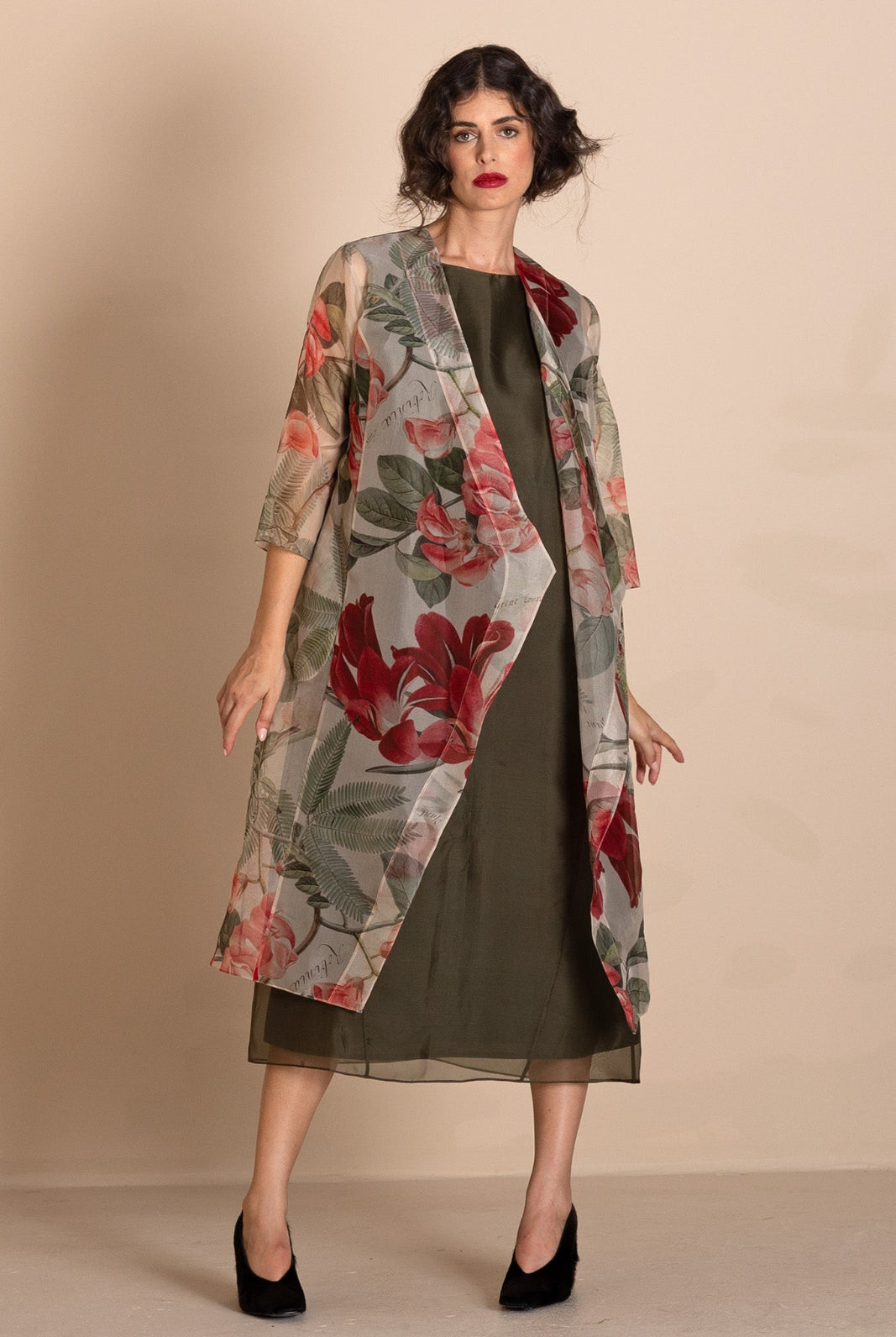 A woman models a sheer, open-front midi duster coat featuring a red and green floral print over a dark olive green dress, set against a plain beige studio background.
