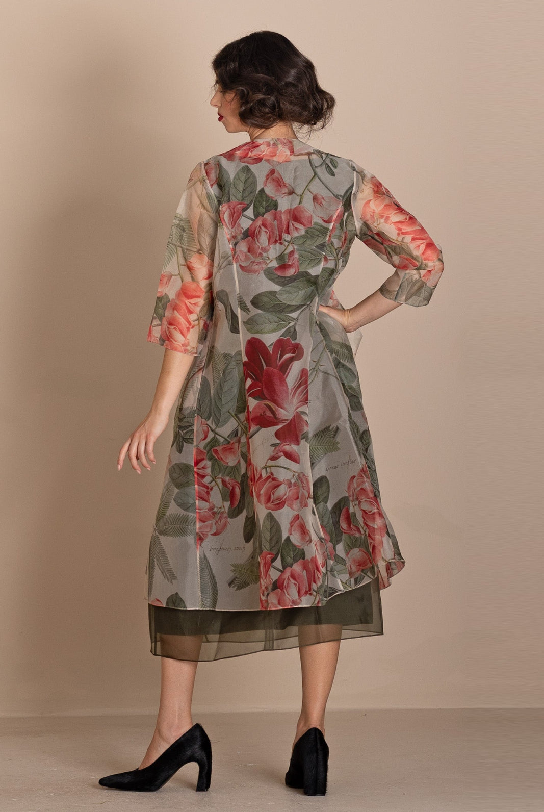 A woman is shown from the back wearing a sheer floral printed tunic dress with three-quarter sleeves, featuring red and pink flowers with green leaves on a gray base, paired with black heels, set against a solid beige studio background.