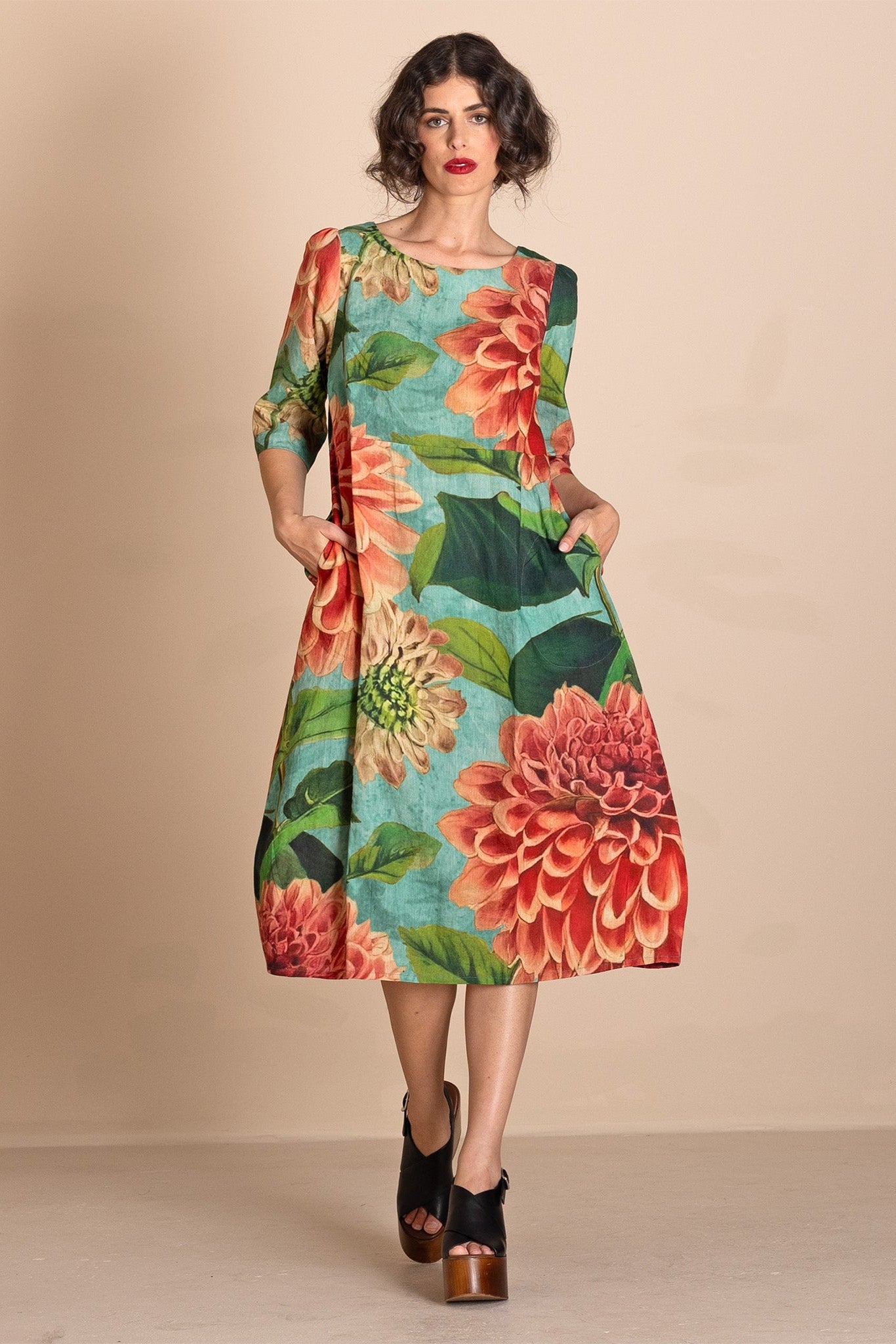 A woman models a knee-length A-line dress with three-quarter sleeves, featuring a vibrant floral print of red and orange flowers with green leaves on a turquoise blue background. She is wearing black platform sandals and posing against a solid beige studio backdrop.