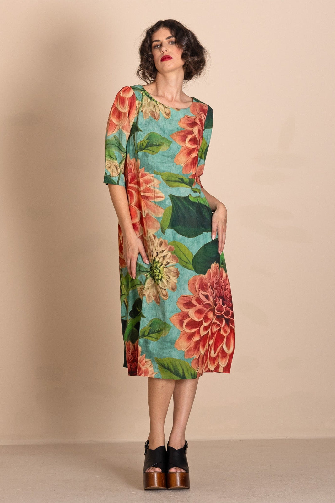 A woman models a knee-length dress with a vibrant floral print featuring large red and orange flowers, green leaves, and a turquoise blue background. She is wearing black platform heels against a solid beige studio backdrop.