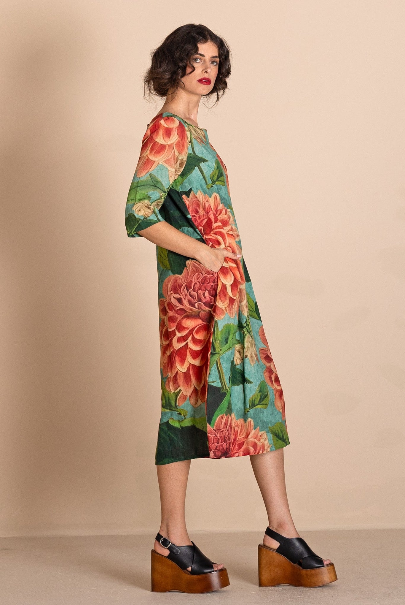 A woman models a knee-length dress with a vibrant green base and a large red and orange floral print, styled with black platform sandals against a solid beige studio background.