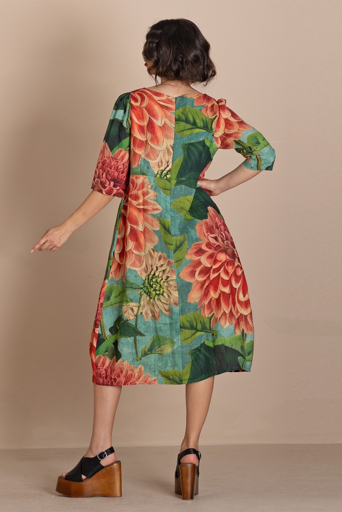 A woman stands with her back to the camera wearing a knee-length dress with a large floral print in red, orange, green, and teal, paired with black wedge sandals, against a solid beige studio background.