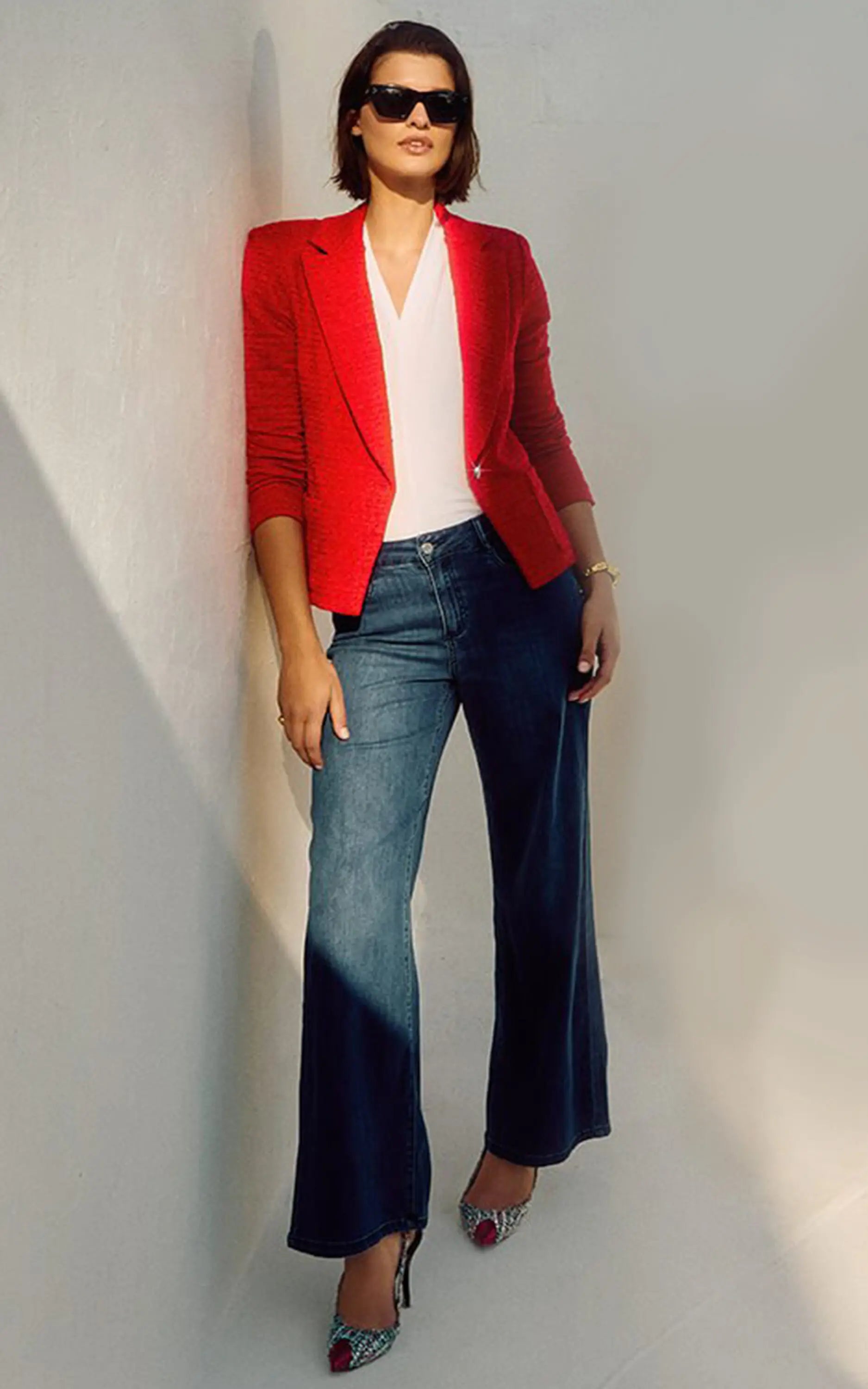 Woman wearing a red blazer, white top, and blue jeans against a plain background