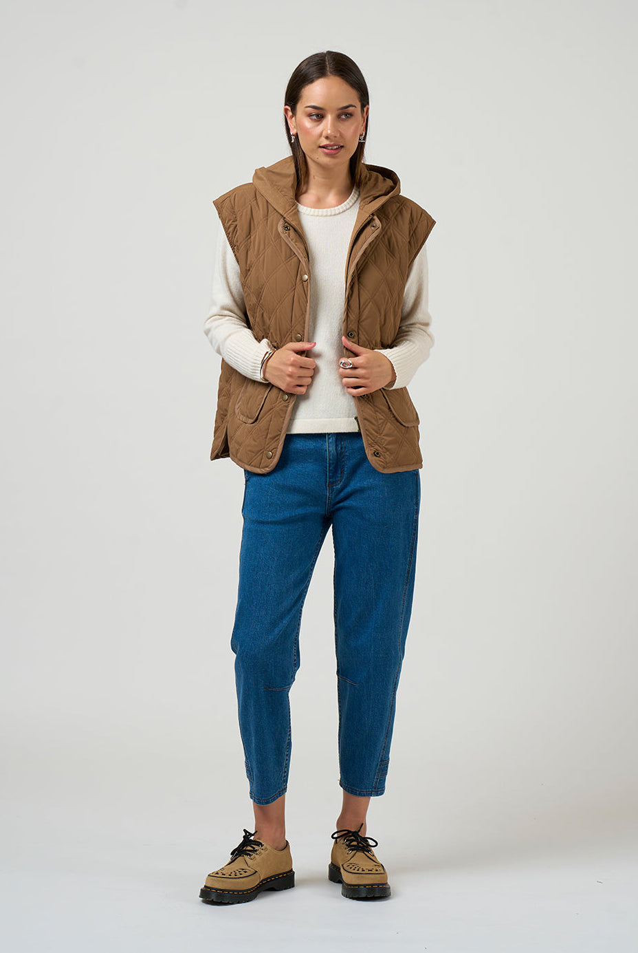 Woman wearing a brown vest, white sweater, blue jeans, and brown shoes on a white background
