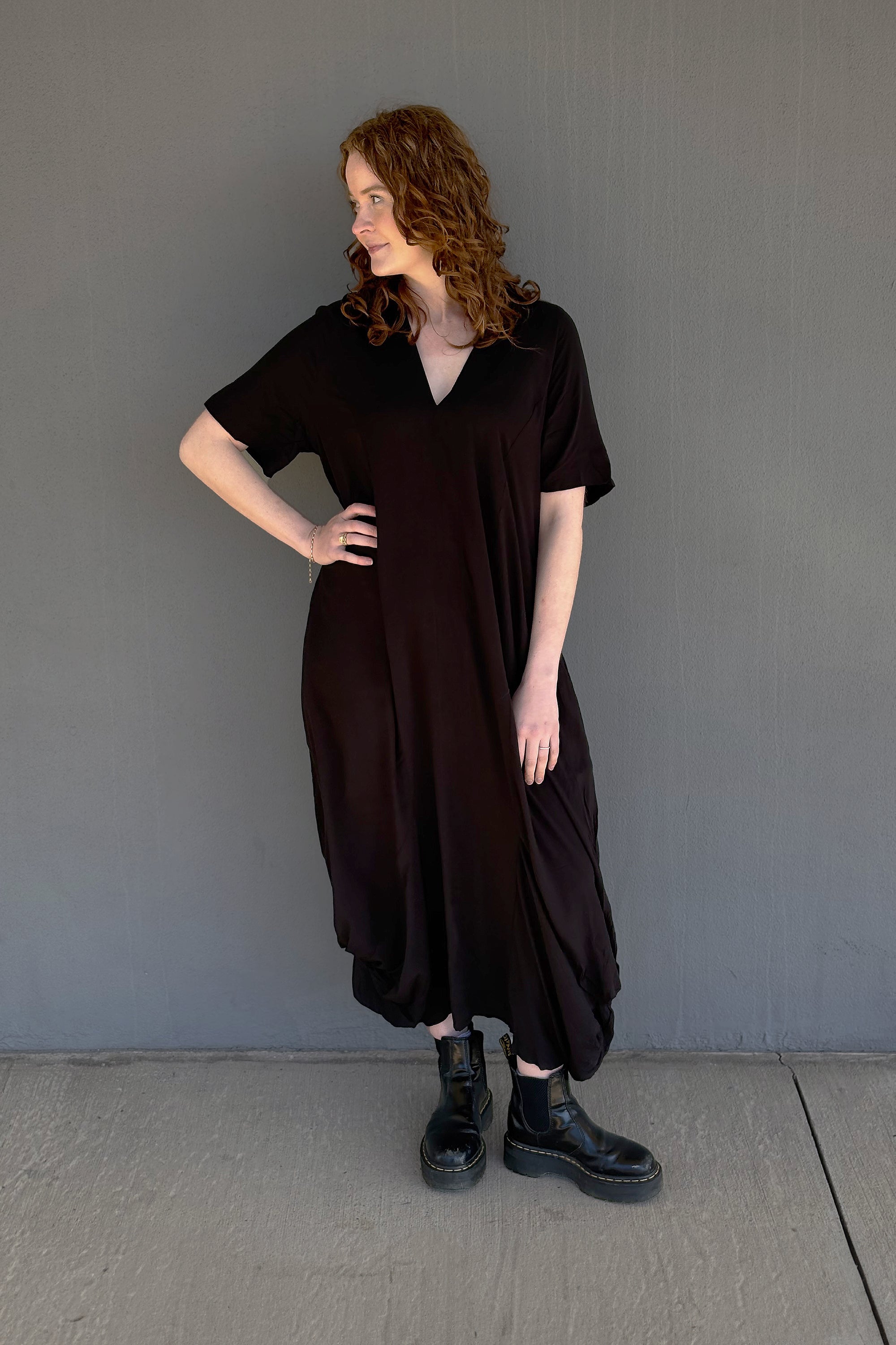 A woman with curly red hair models a loose, dark brown short-sleeve midi dress with a V-neckline, paired with black combat boots, standing against a plain gray wall.
