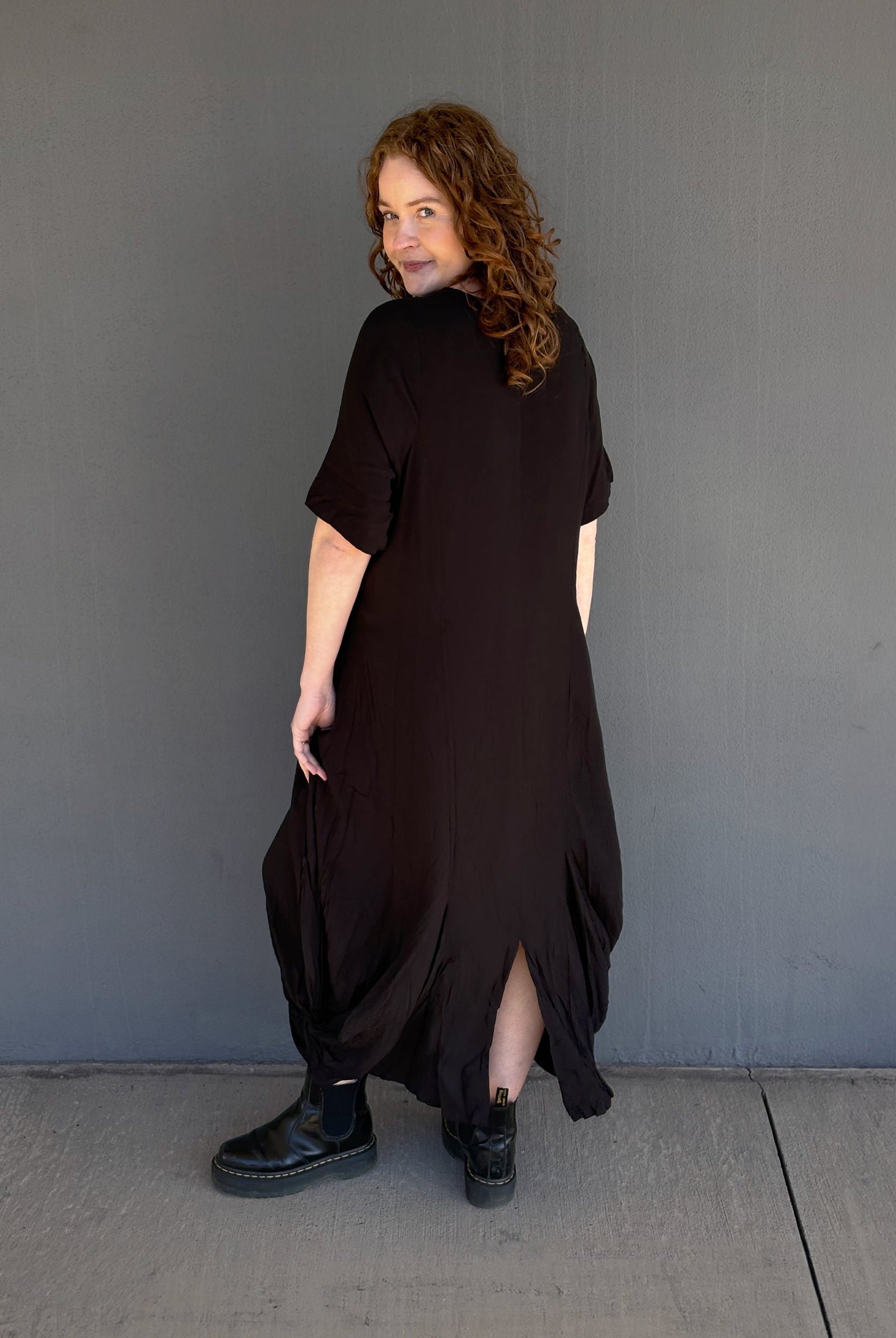 A woman with curly brown hair is shown from the back wearing a loose, oversized black dress with short sleeves and black boots, standing against a plain gray wall.