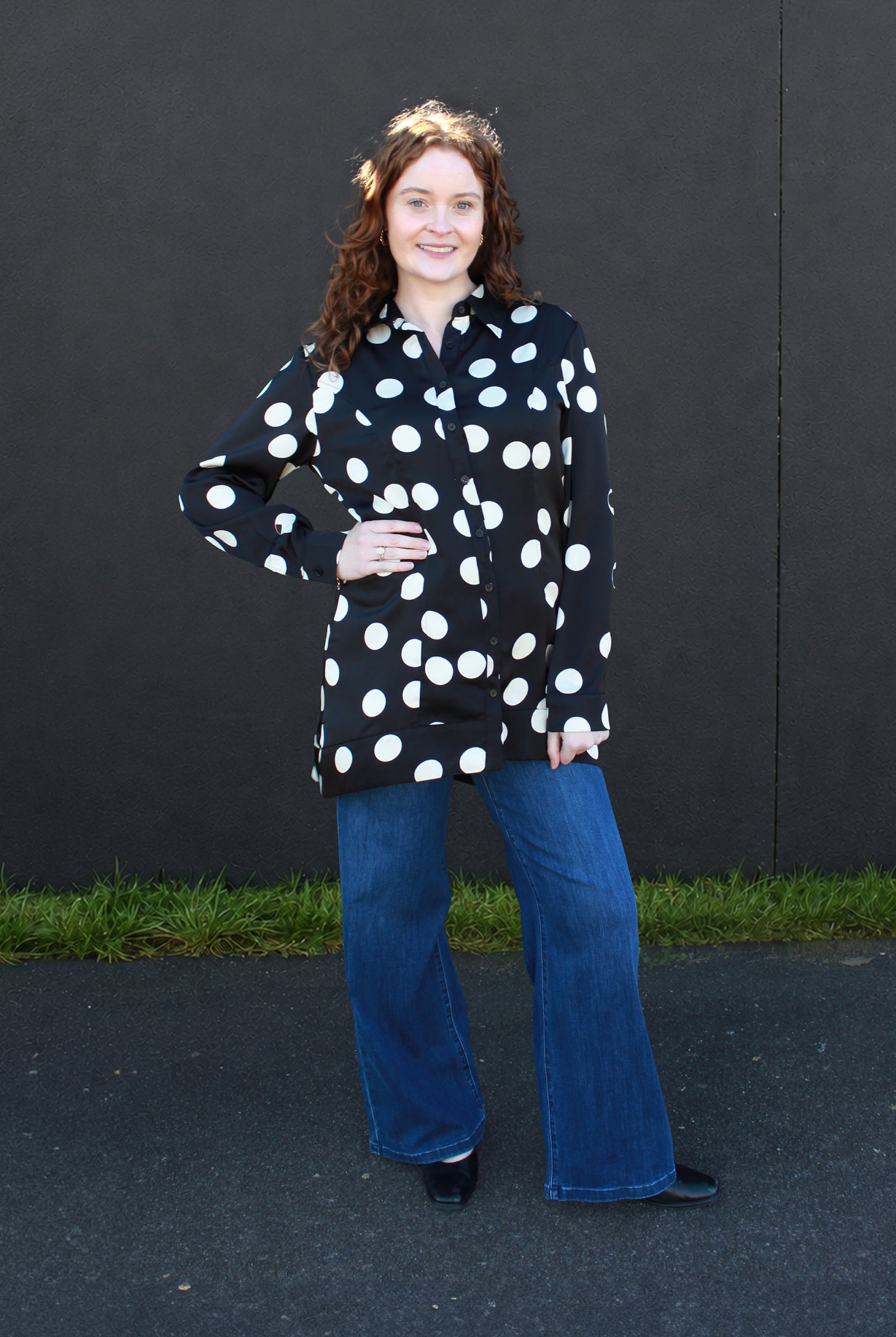 A woman with curly brown hair stands outdoors against a dark wall, wearing a black long-sleeve button-up shirt with large white polka dots and blue wide-leg jeans.