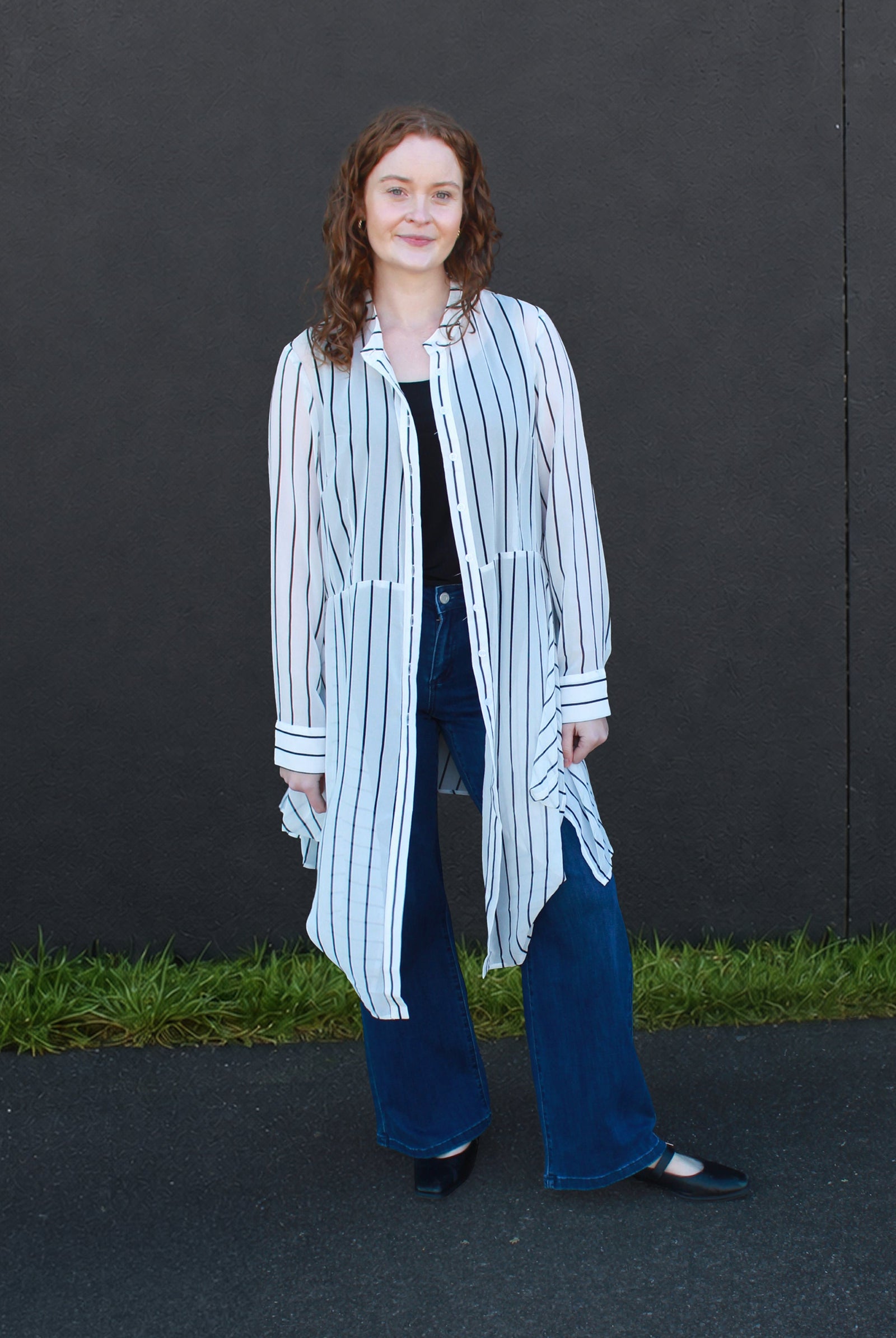 A woman stands outdoors against a dark gray wall wearing a long-sleeve white shirt with thin vertical black stripes, a black top underneath, wide-leg blue jeans, and black flats.