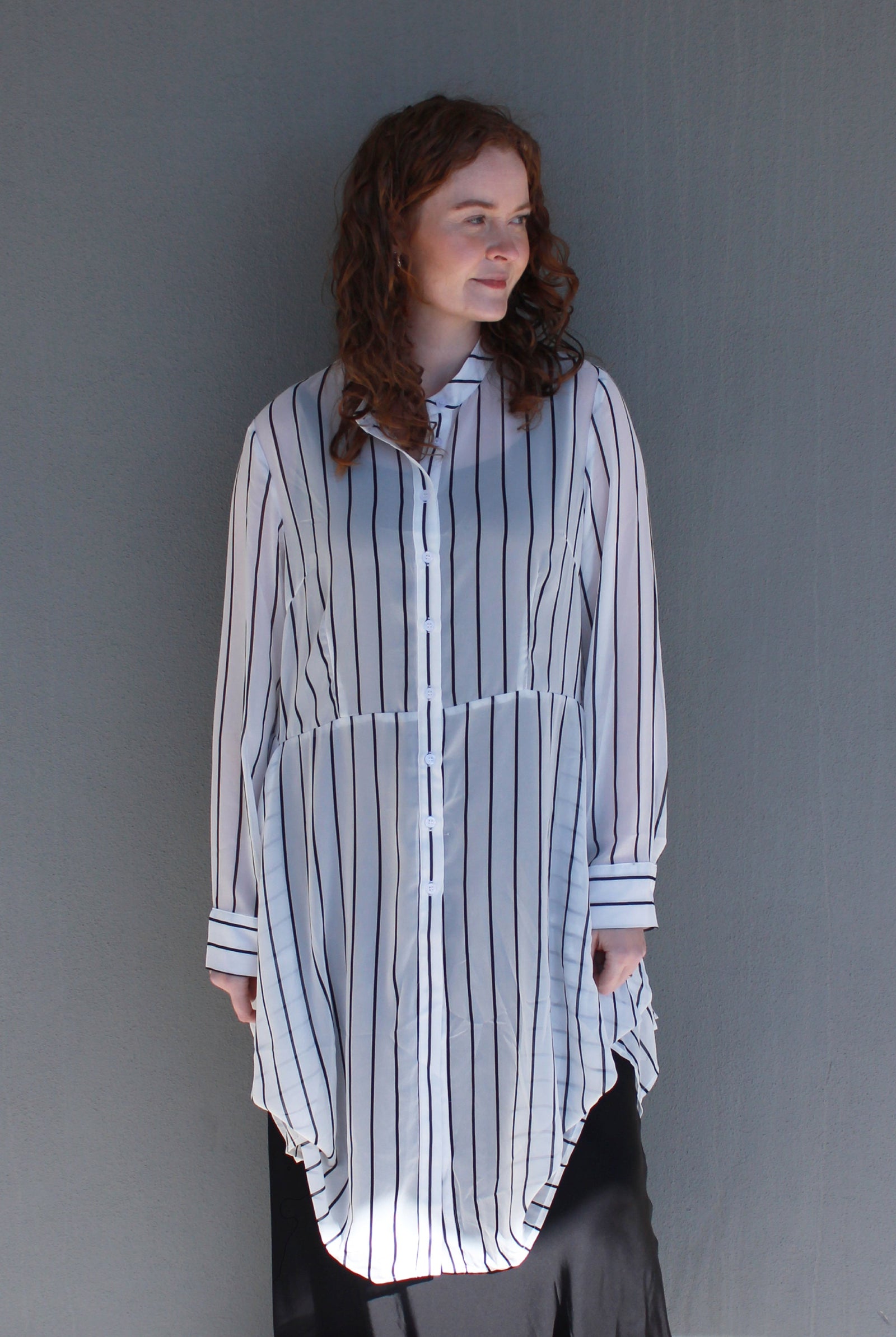 A woman with curly brown hair wearing a white button-up shirt with thin black vertical stripes and a high-low hem, paired with black bottoms, standing against a plain gray wall.