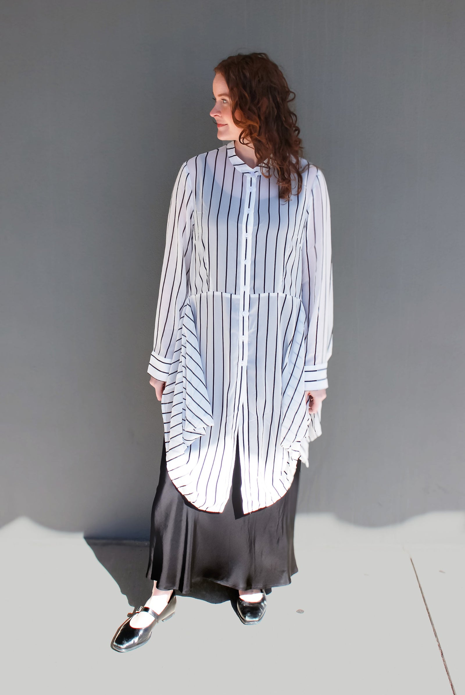 A woman stands against a plain gray wall wearing a loose white button-front shirt with thin vertical navy stripes and a long dark skirt. She is photographed in bright sunlight with a hard shadow on the ground.