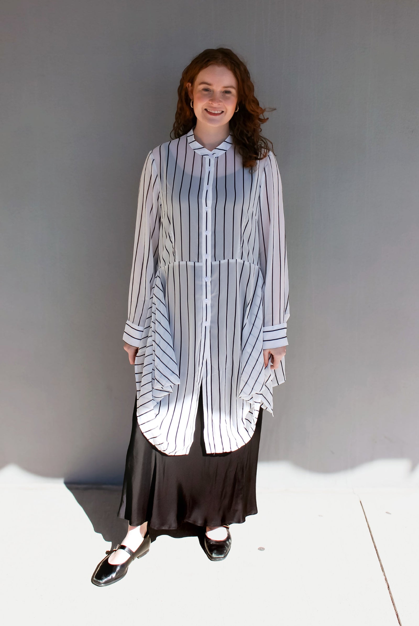 A woman modeling a long-sleeve button-front shirt dress with vertical blue and white stripes, worn over a black maxi skirt and black Mary Jane shoes, standing against a gray wall.