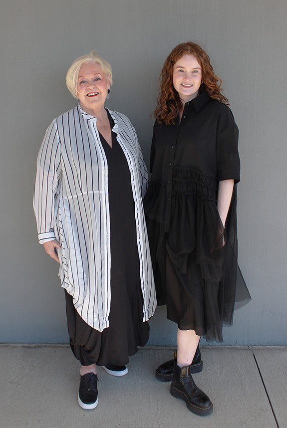 Two women standing side-by-side outdoors against a gray wall. The woman on the left has short blonde hair and wears a white button-up shirt with thin black vertical stripes over a black dress and black sneakers.