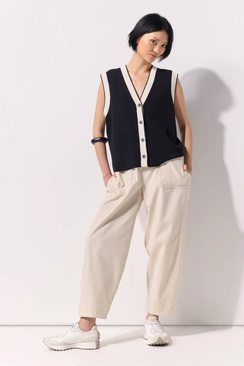Woman wearing a black buttoned up sleeveless knit vest with white contrast trim and black buttons against a white wall.