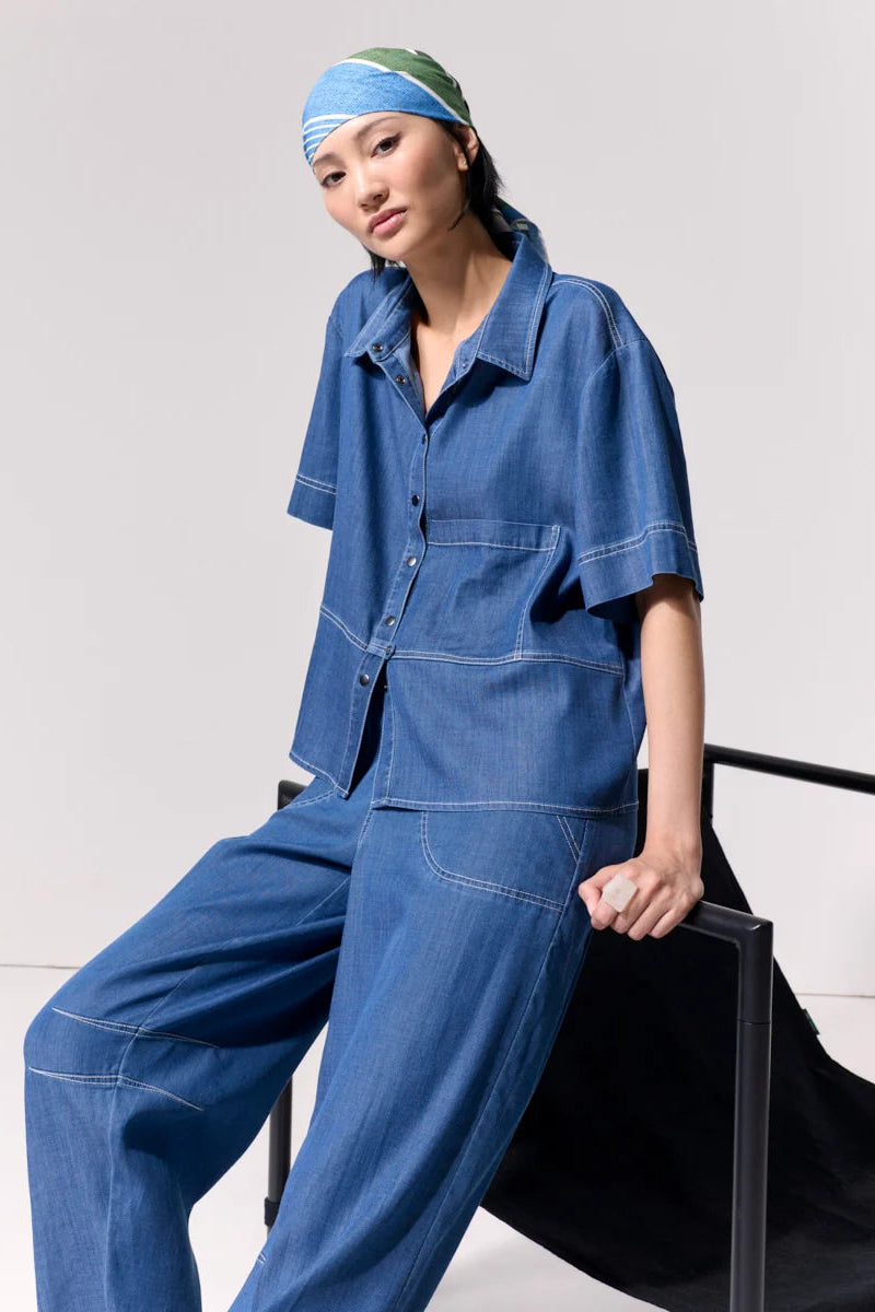 Woman wearing a short sleeve light denim shirt with collar and front left side pocket with contrast stitching against a white background.
