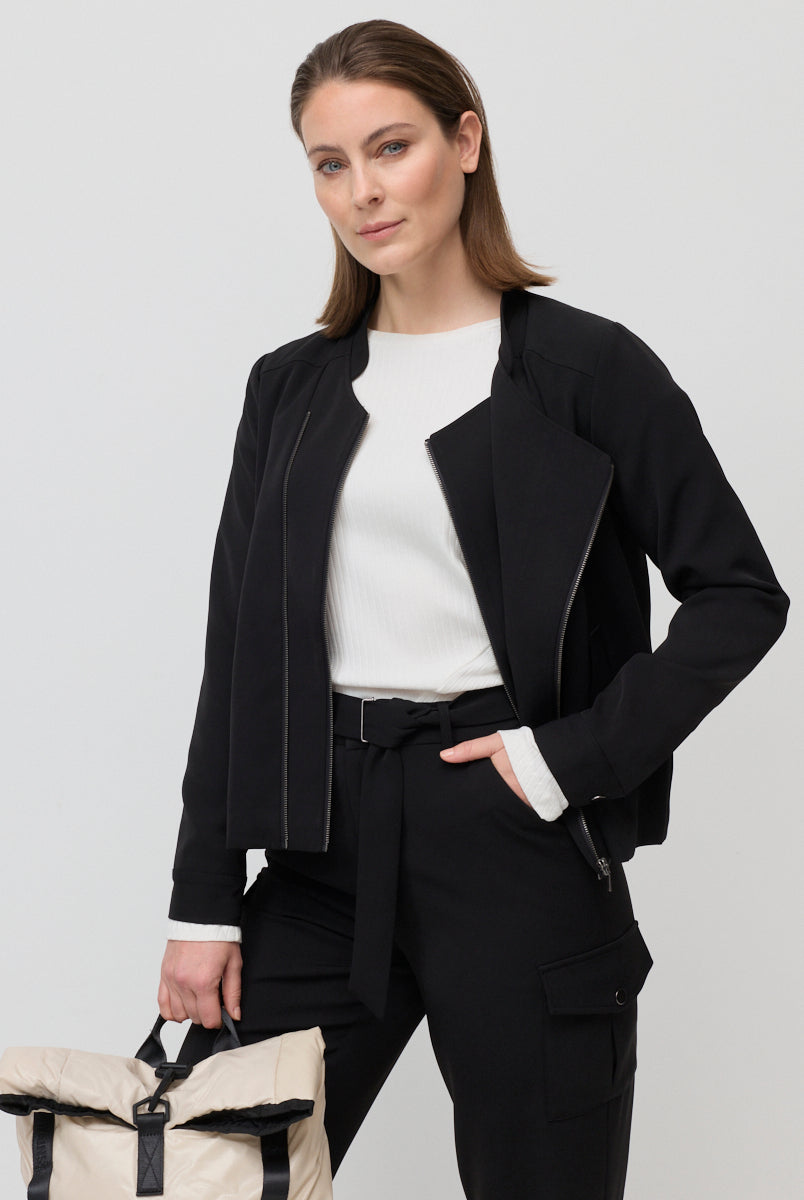 Woman wearing a black jacket and pants with a white top, holding a beige backpack on a light gray background.