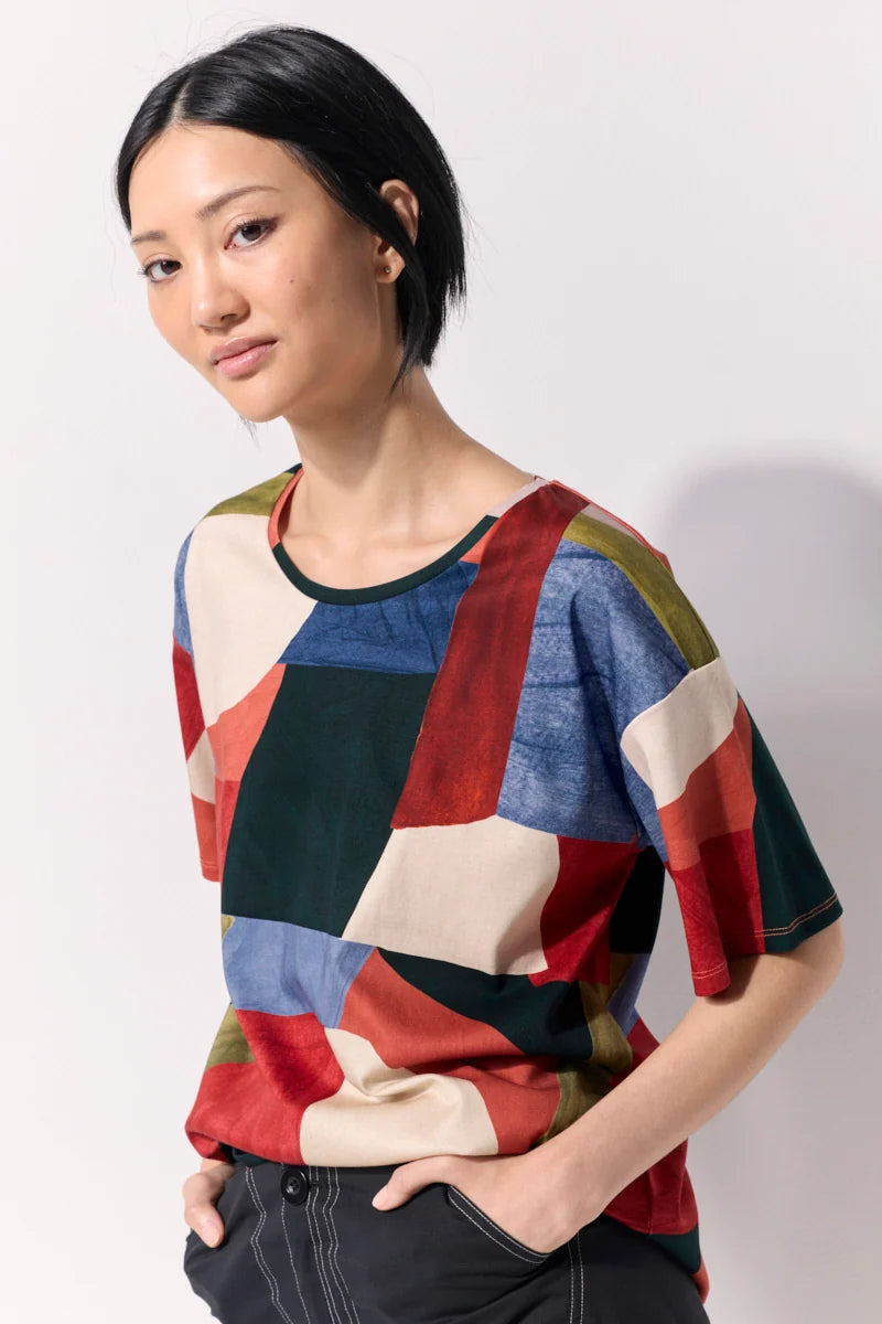 A woman wearing a short-sleeve crewneck top with a multicolor geometric patchwork pattern, paired with dark gray pants, photographed against a plain light gray studio background.