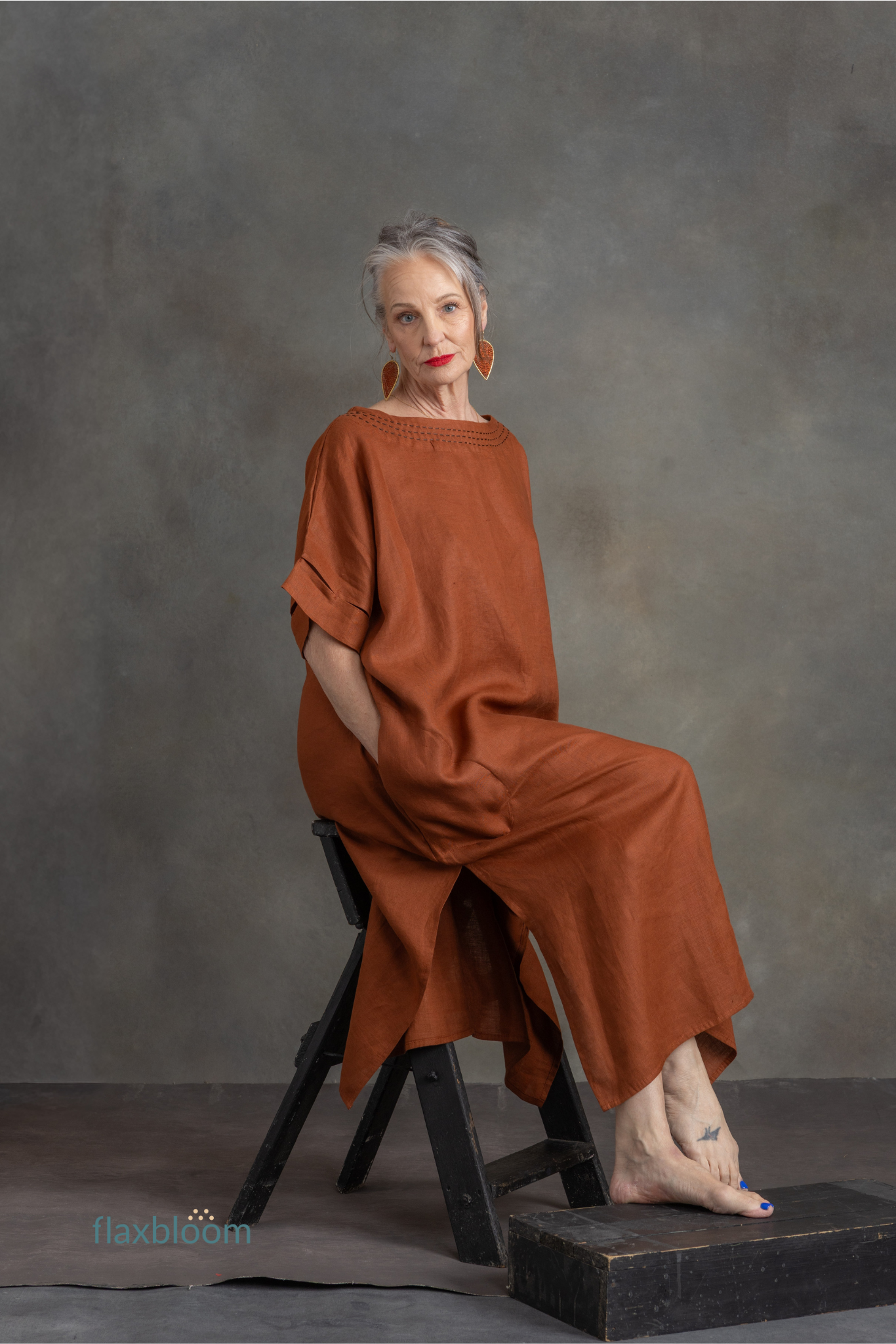 Woman in a rust-colored kaftan sitting on a black stool against a gray background