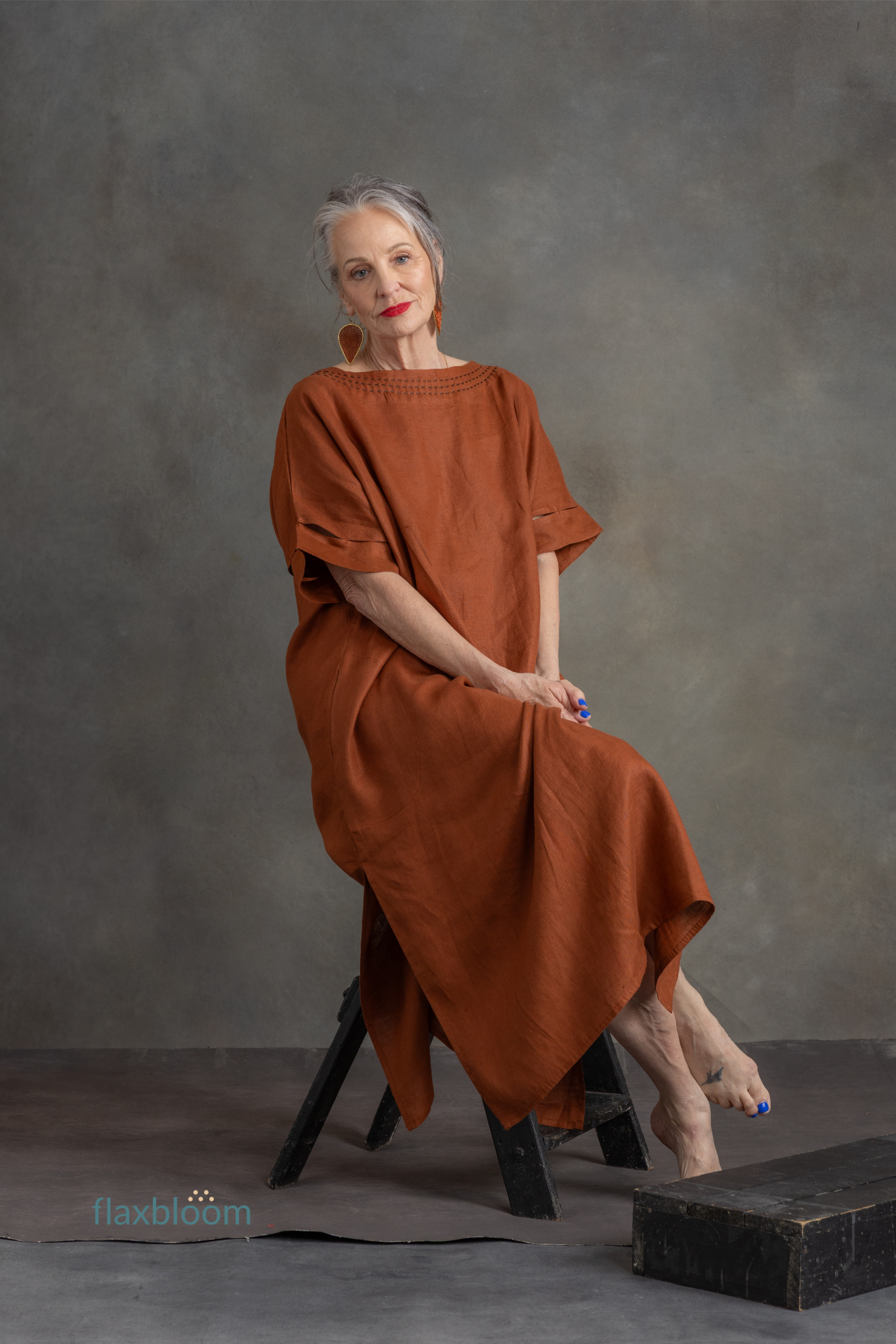 Woman in a rust-colored kaftan sitting on a black stool against a gray background