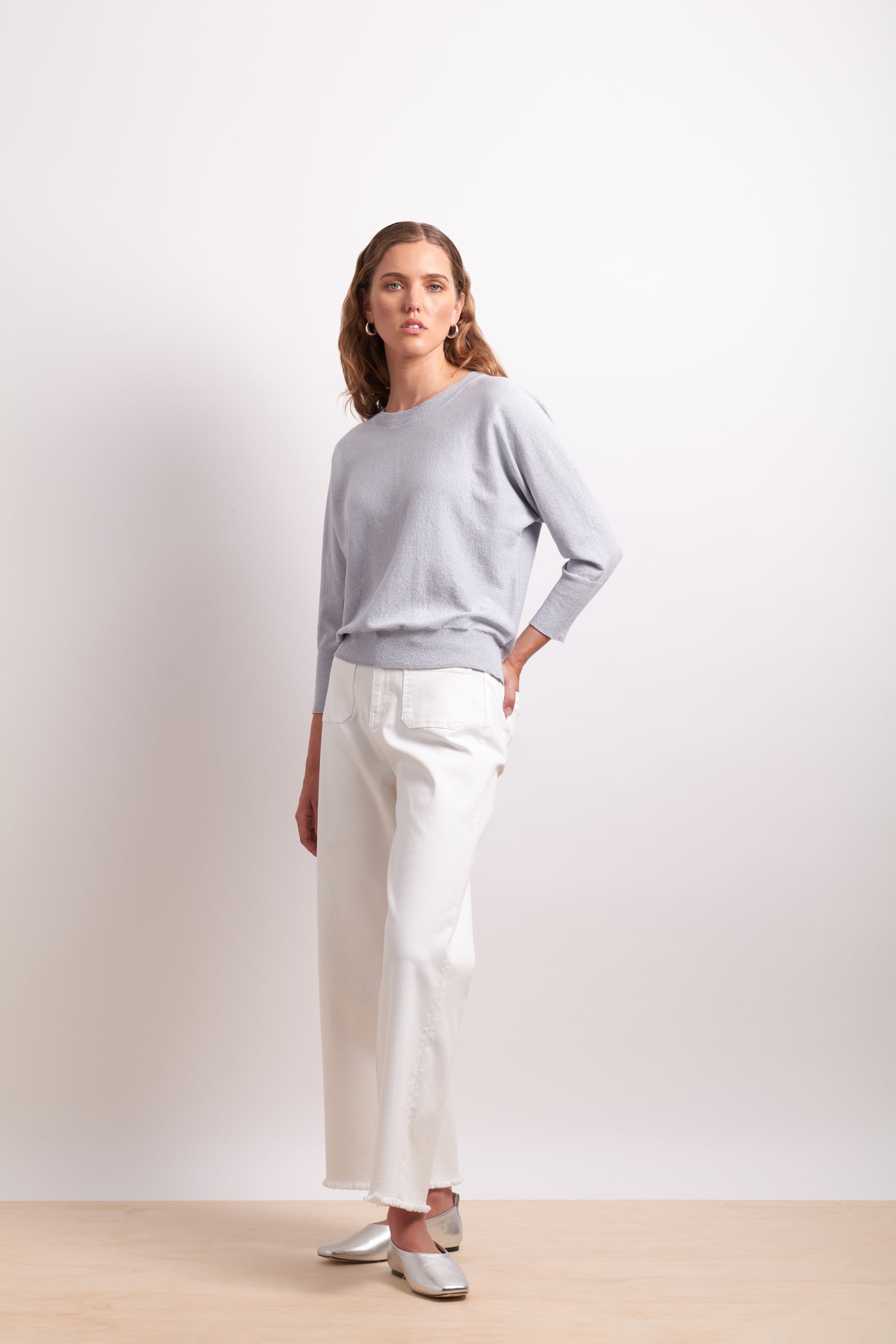 Woman wearing a light gray sweater and white pants on a plain background