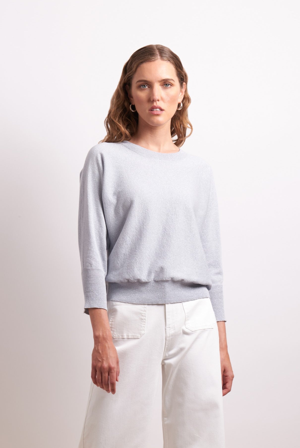 Woman wearing a light gray sweater and white pants against a plain background