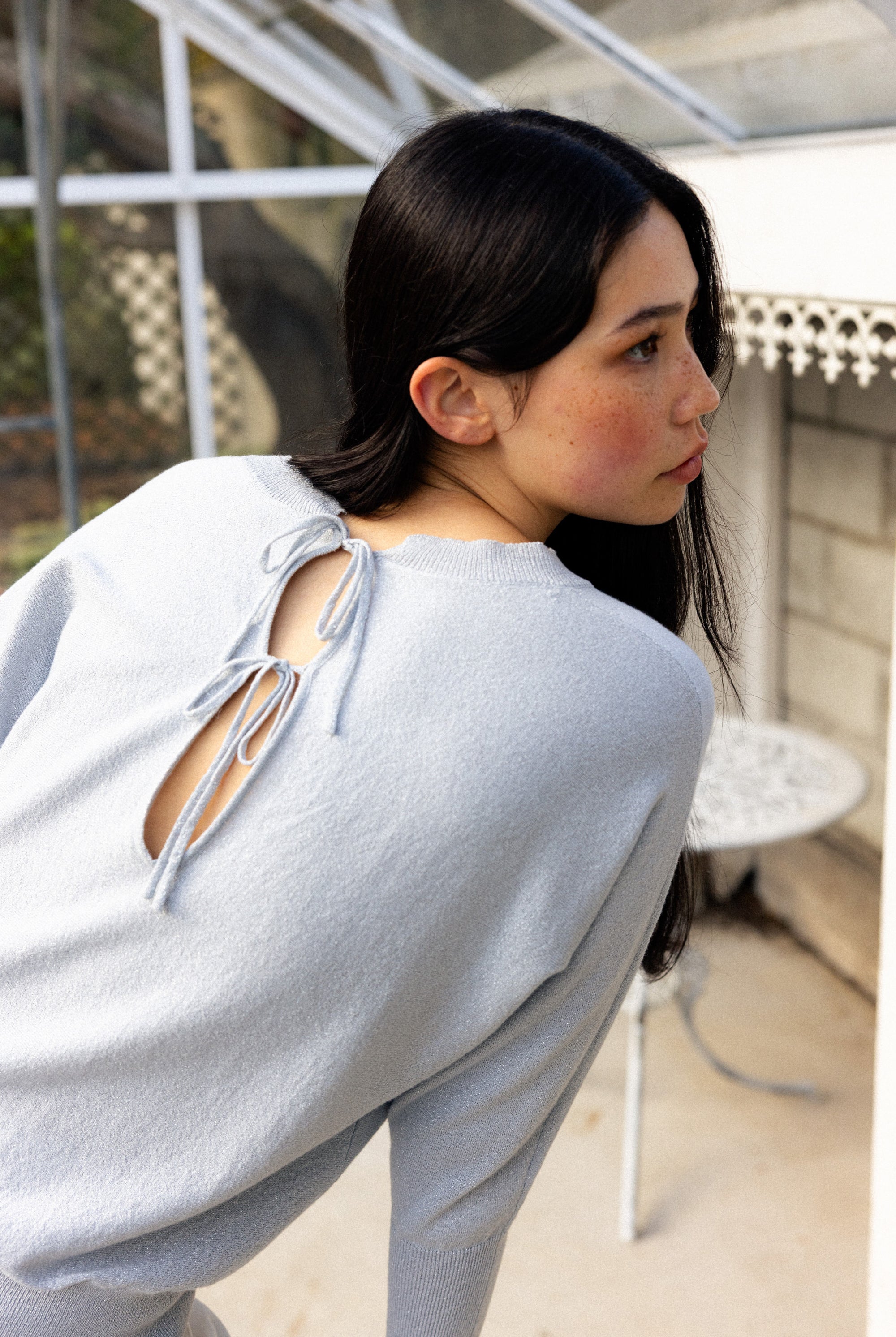 Woman wearing a light gray sweater with cut-out back in an indoor setting.