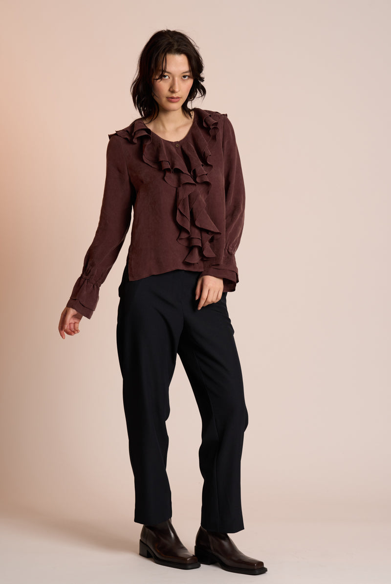 Woman wearing a brown ruffled blouse and black pants on a beige background