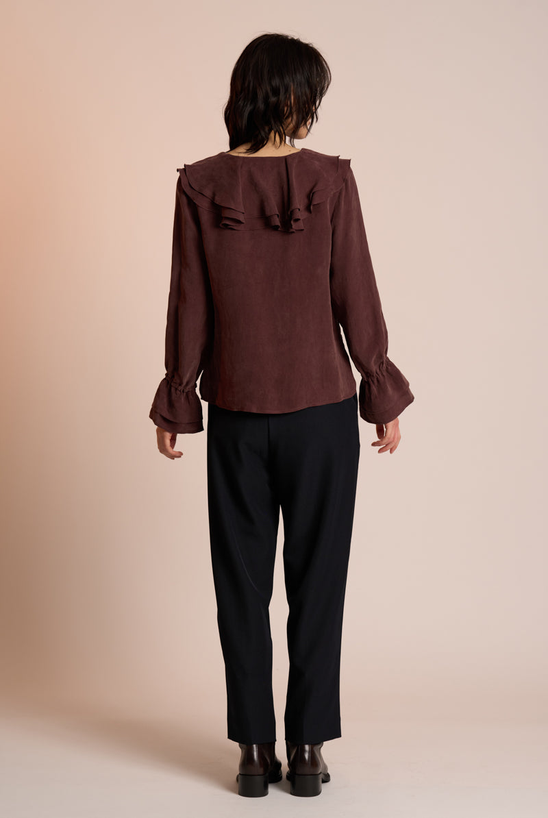 Person wearing a brown blouse with ruffled details and black pants on a beige background back view